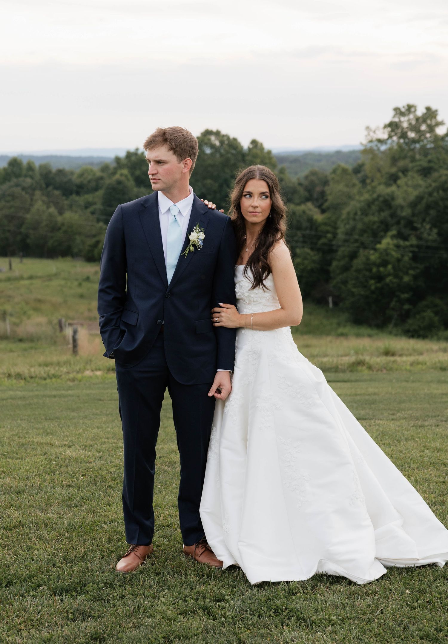 Bride in white gown and groom in navy suit pose together on green lawn with scenic countryside views.