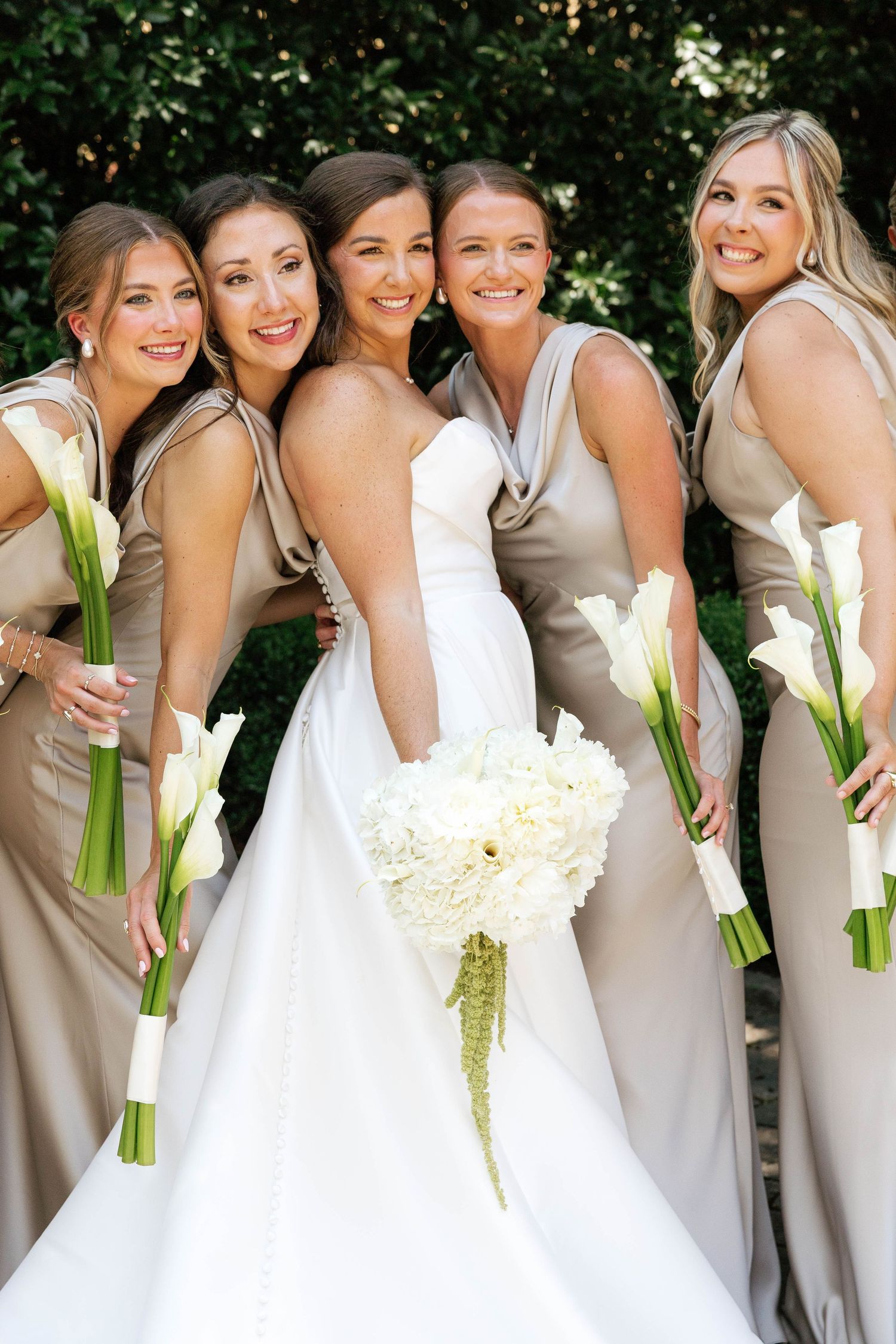Bride in white strapless gown poses with four bridesmaids in taupe dresses holding calla lily bouquets.