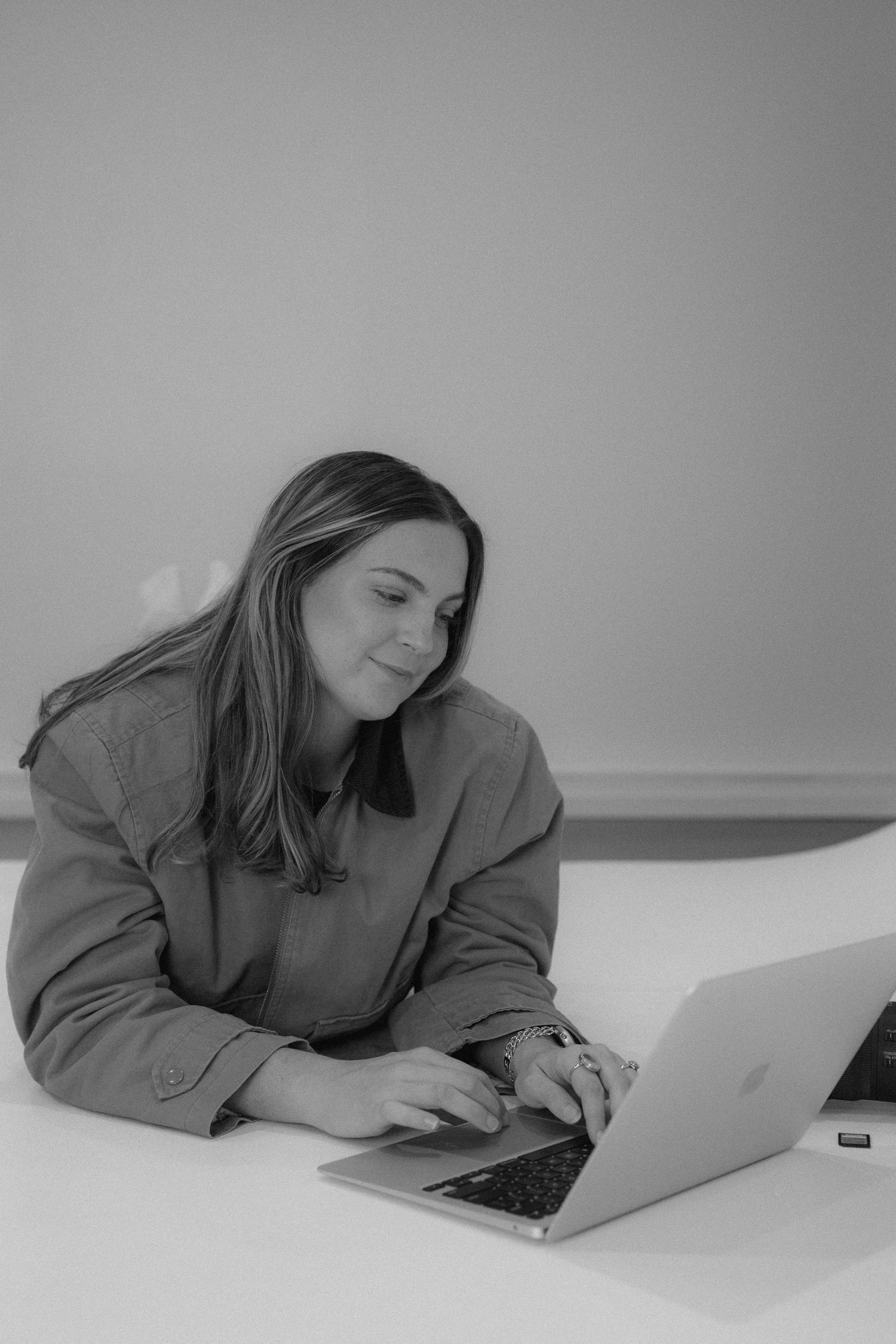 Black and white photo of a smiling woman working on a MacBook laptop at a white desk.