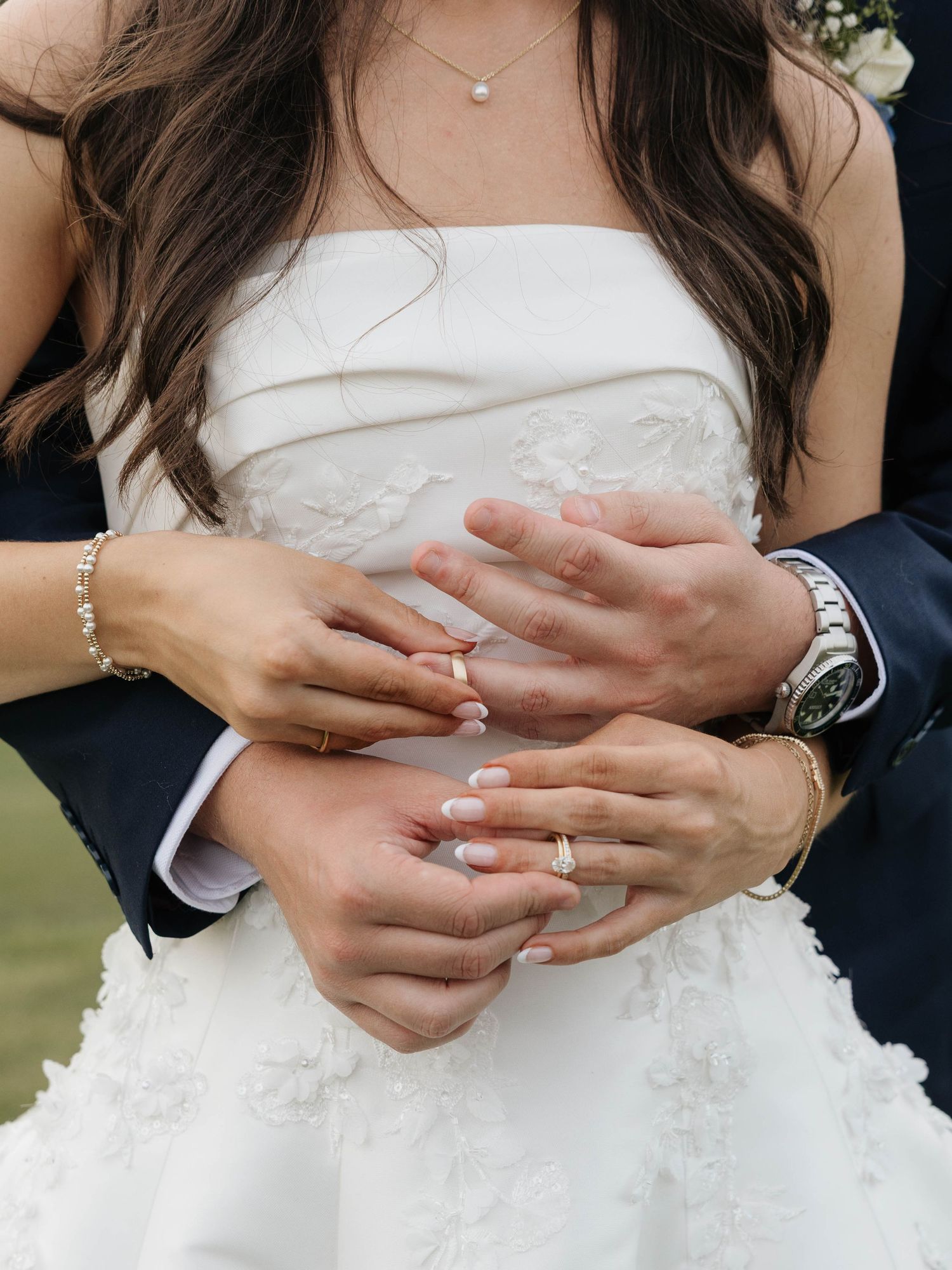 Close-up of bride and groom hands showing wedding rings, bride wearing white lace strapless gown.