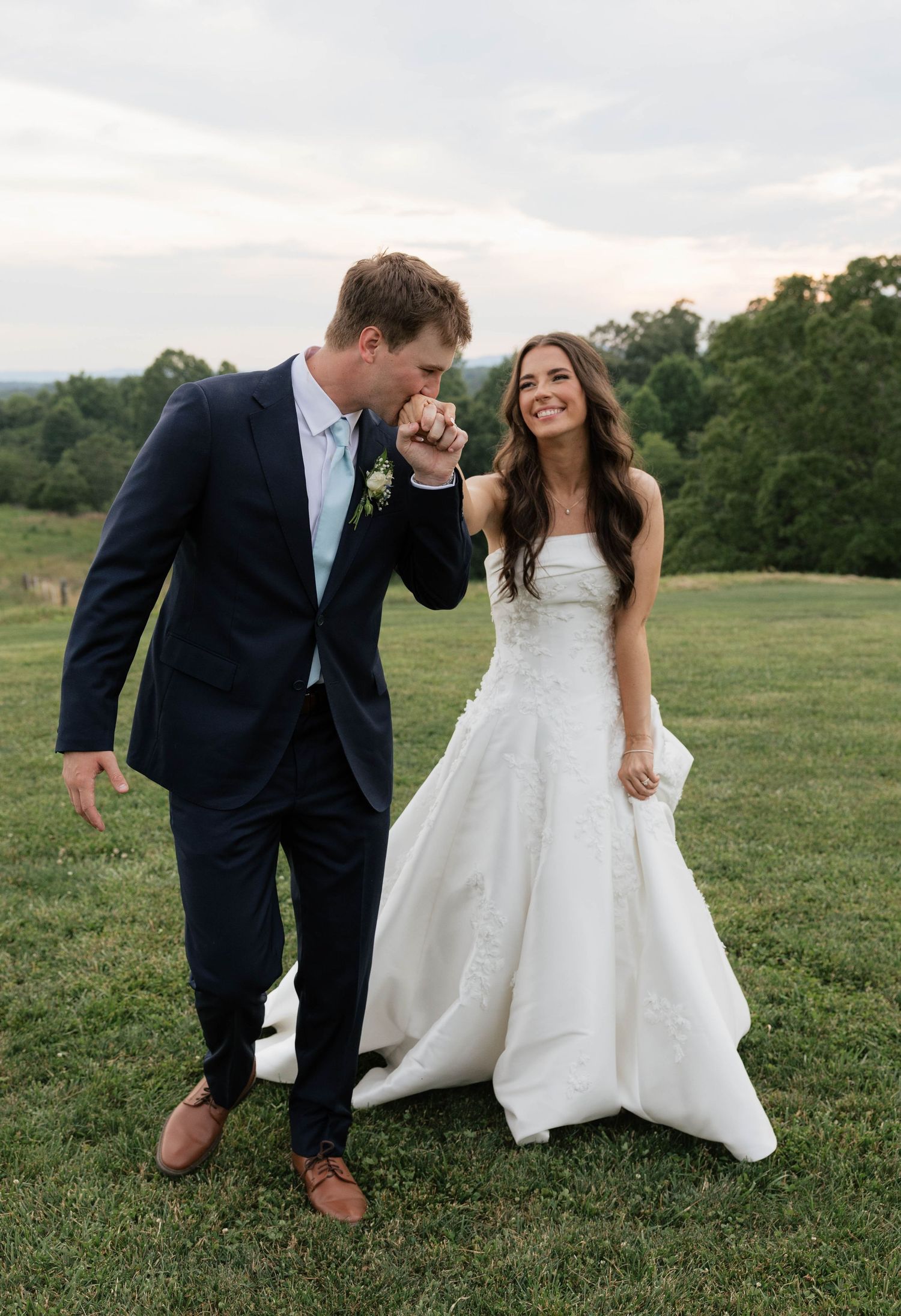 Groom in navy suit kisses bride's hand as she laughs in white strapless gown on a grassy hillside.