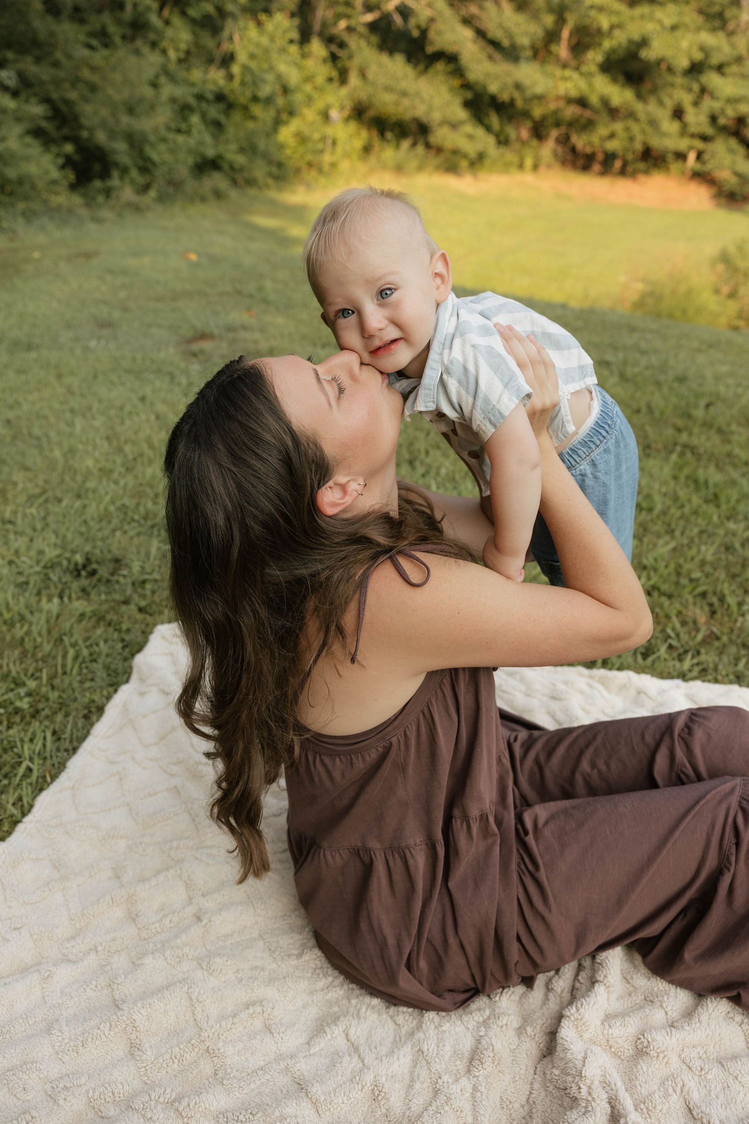 A young mother in a brown dress lies on a blanket outdoors, lifting and kissing her smiling baby boy.