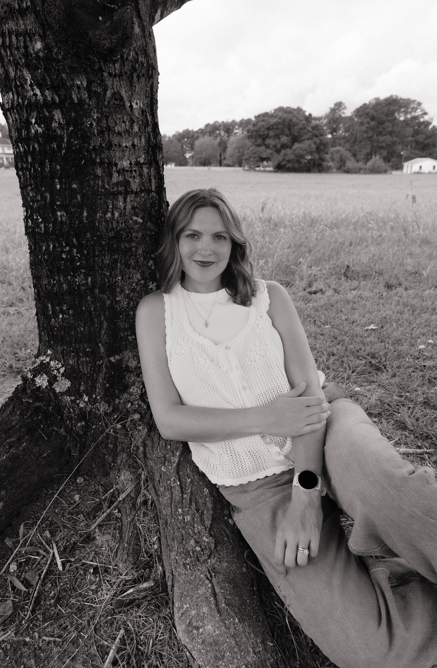 Woman in white sleeveless top leaning against tree in black and white rural landscape photo.