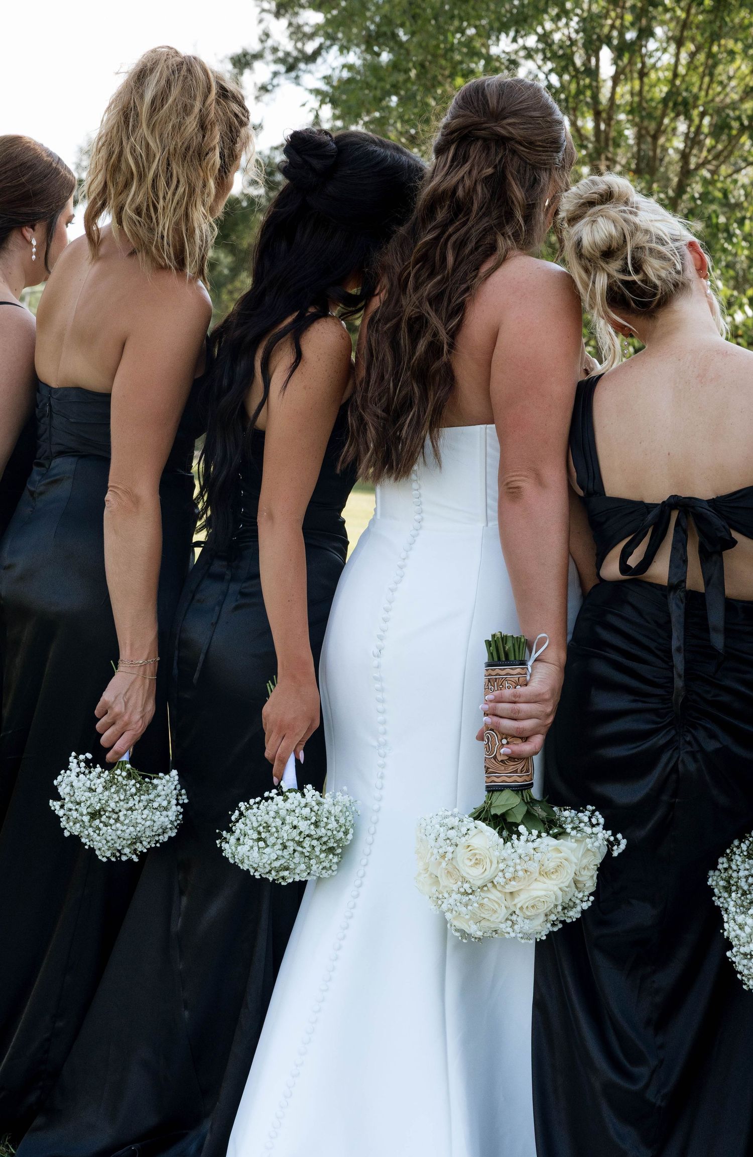 Bride in white gown with bridesmaids in black dresses holding white bouquets, viewed from behind outdoors.