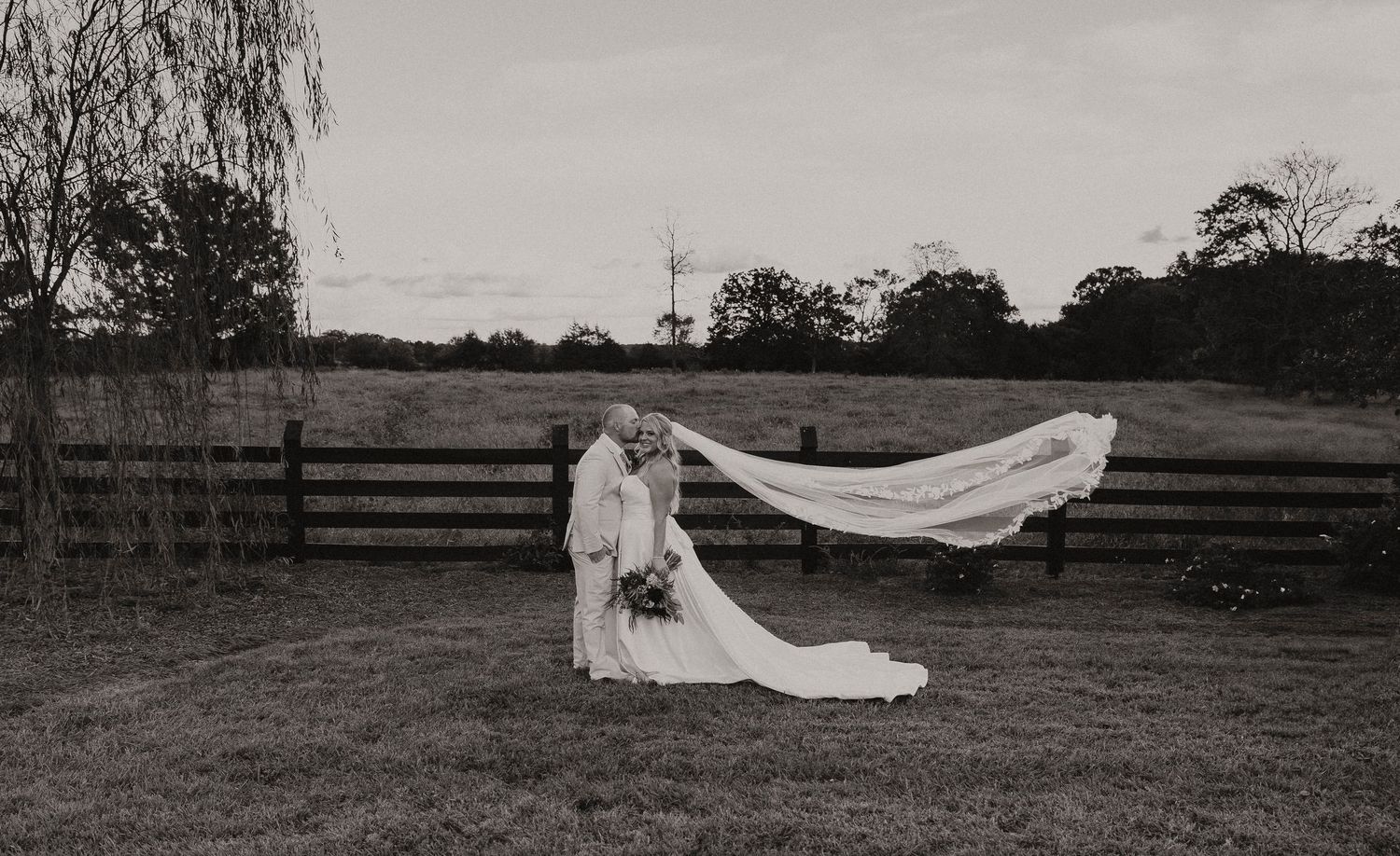 Black and white photo of bride and groom kissing as wedding veil flows in wind by rustic fence.