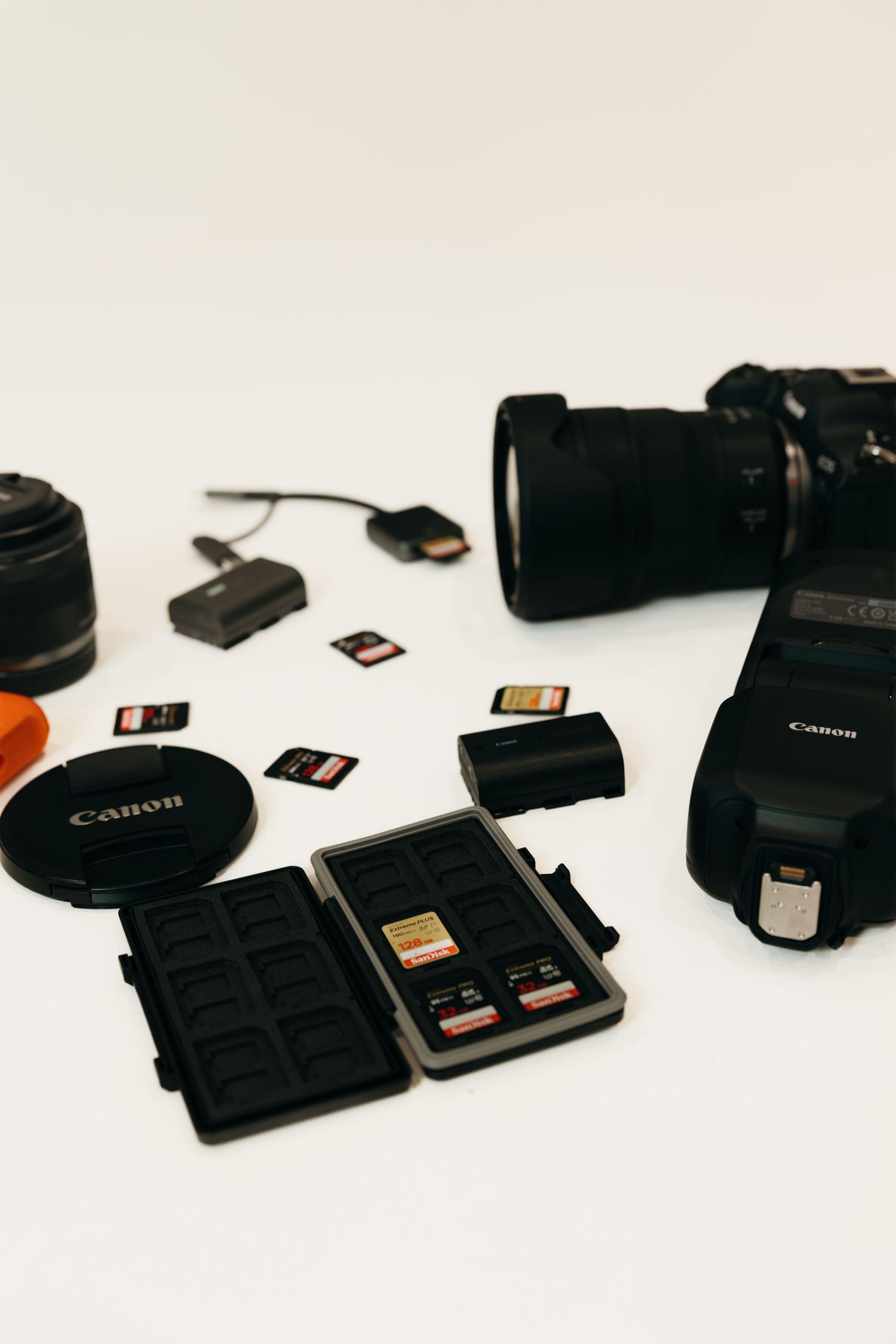Canon camera gear laid out on white surface with SD card case, lenses, battery, and memory card reader.