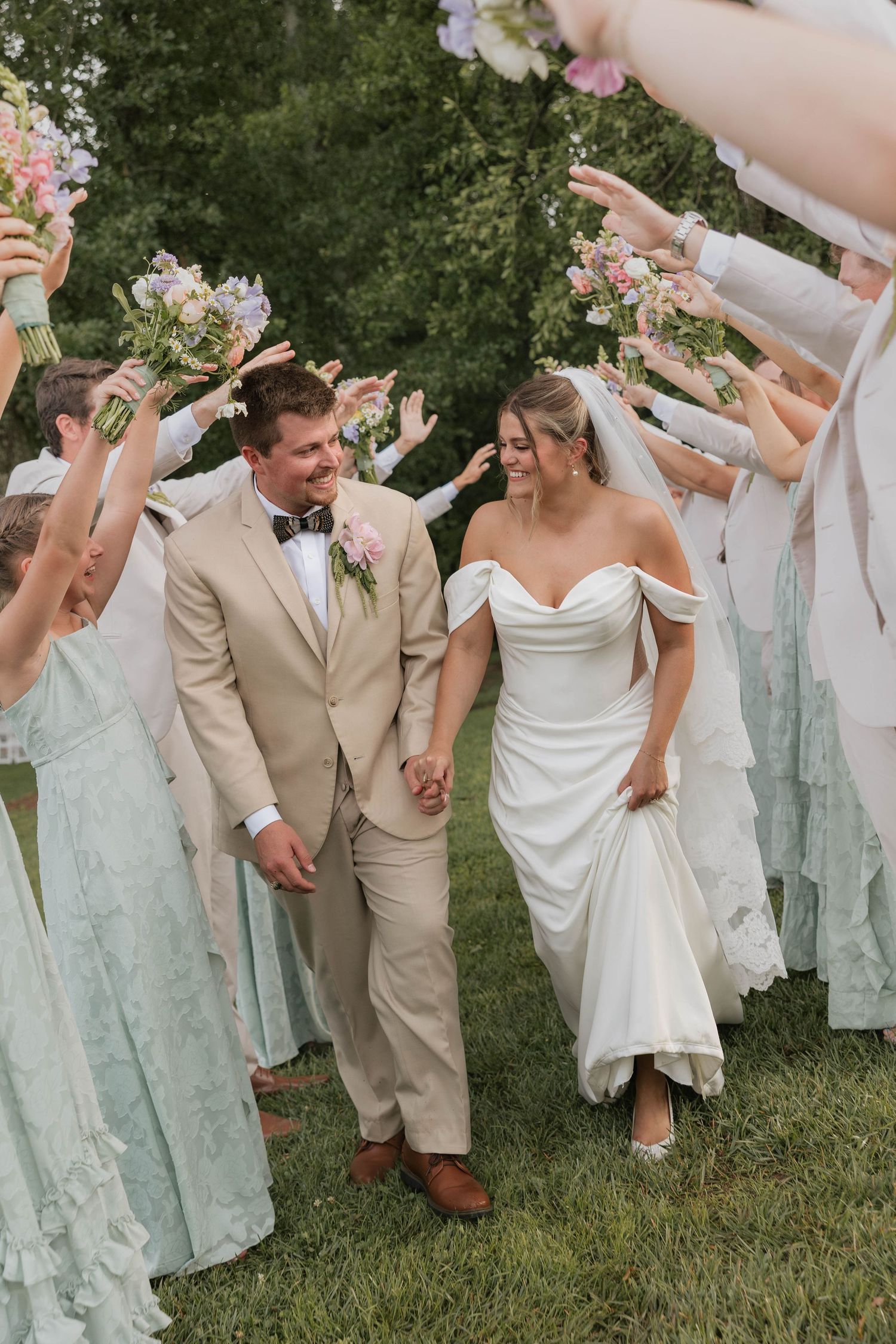 Smiling newlyweds walk through arch of raised bouquets, groom in tan suit, bride in off-shoulder gown.