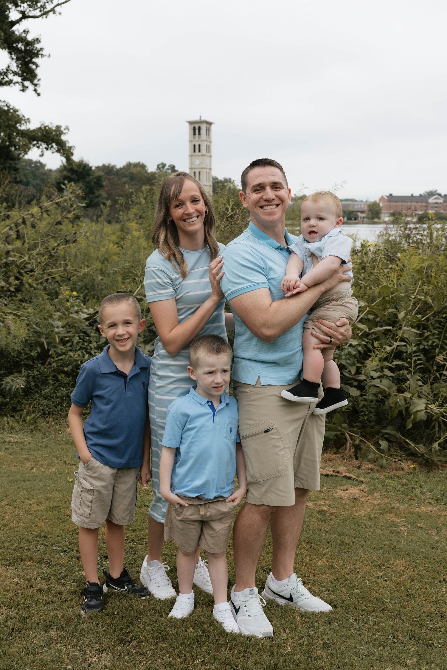 Happy family of five posing outdoors on a green lawn with a church tower visible in the background.