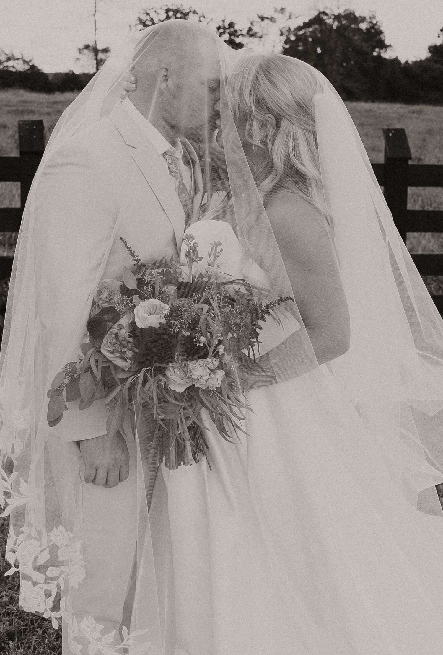Bride and groom share tender kiss beneath flowing veil holding bouquet in black and white photo.