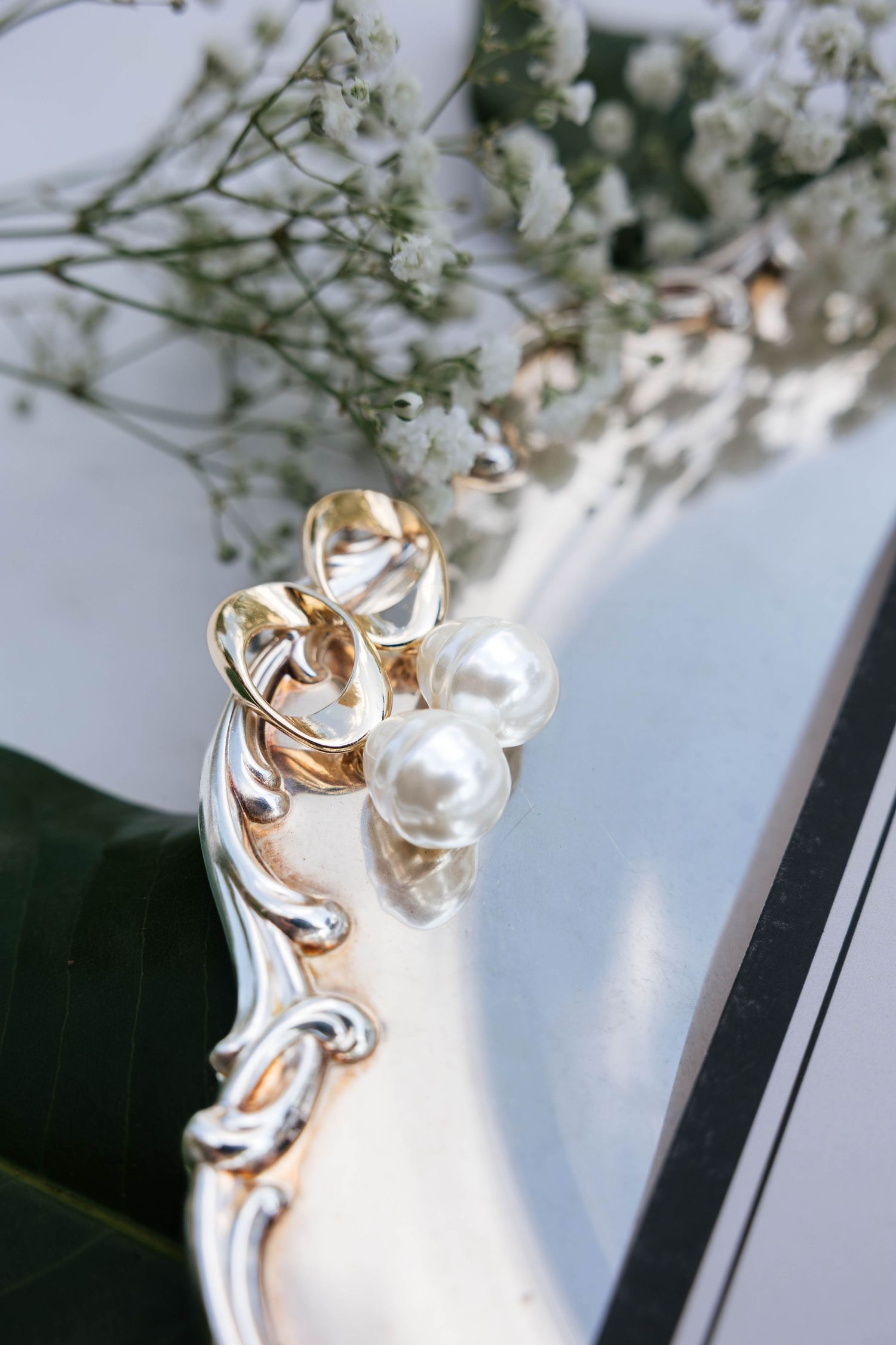 Gold and pearl earring on ornate silver mirror frame with white gypsophila flowers in background.