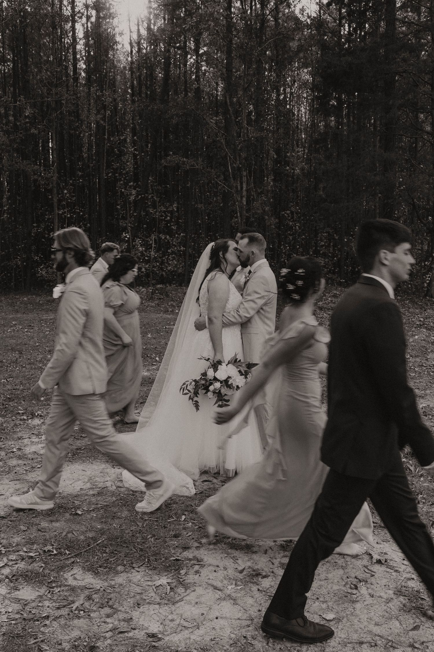 Bride and groom kiss while wedding party walks past in candid black and white forest photo.