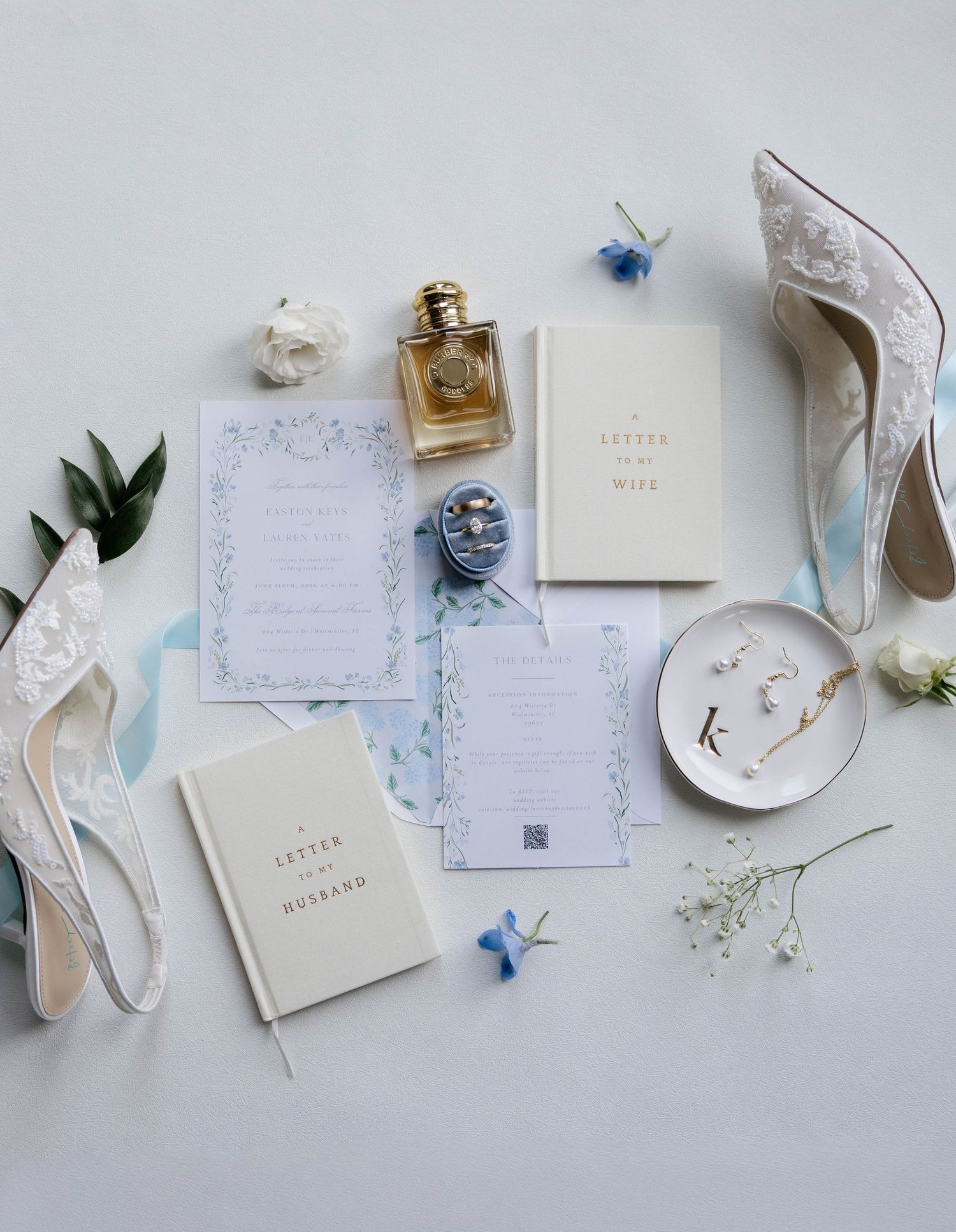 Elegant wedding flat lay with blue floral invitations, perfume, lace heels, rings, and letter books on white background.