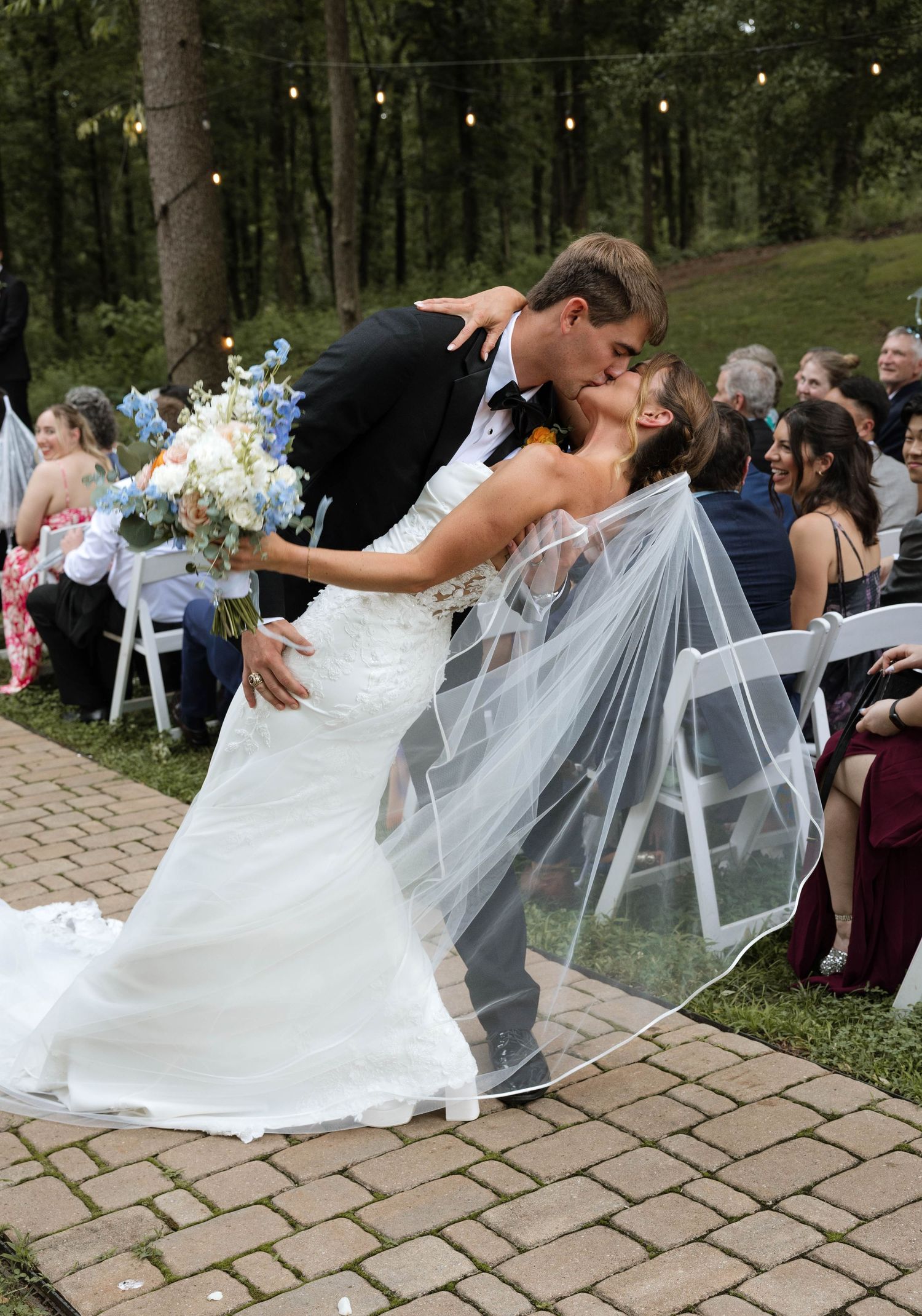 Groom dips bride for romantic kiss along brick aisle at outdoor wedding while guests cheer.