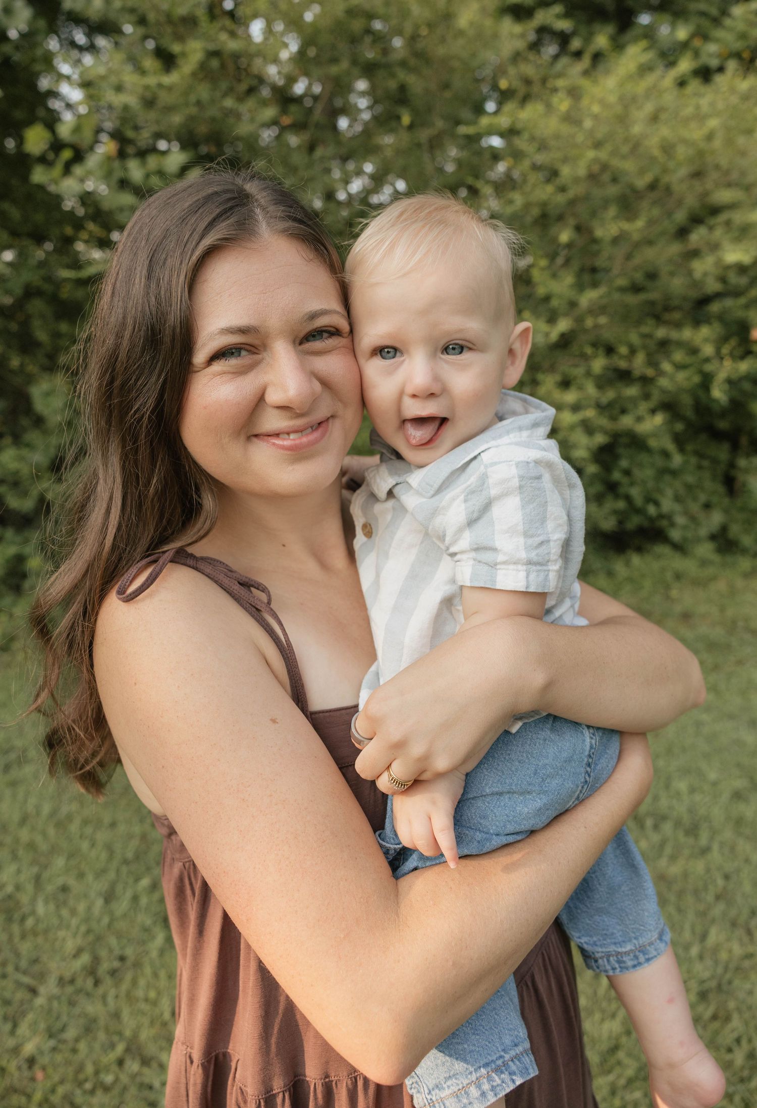 A smiling young woman in a brown dress holds a happy laughing toddler boy outdoors with green trees behind them.