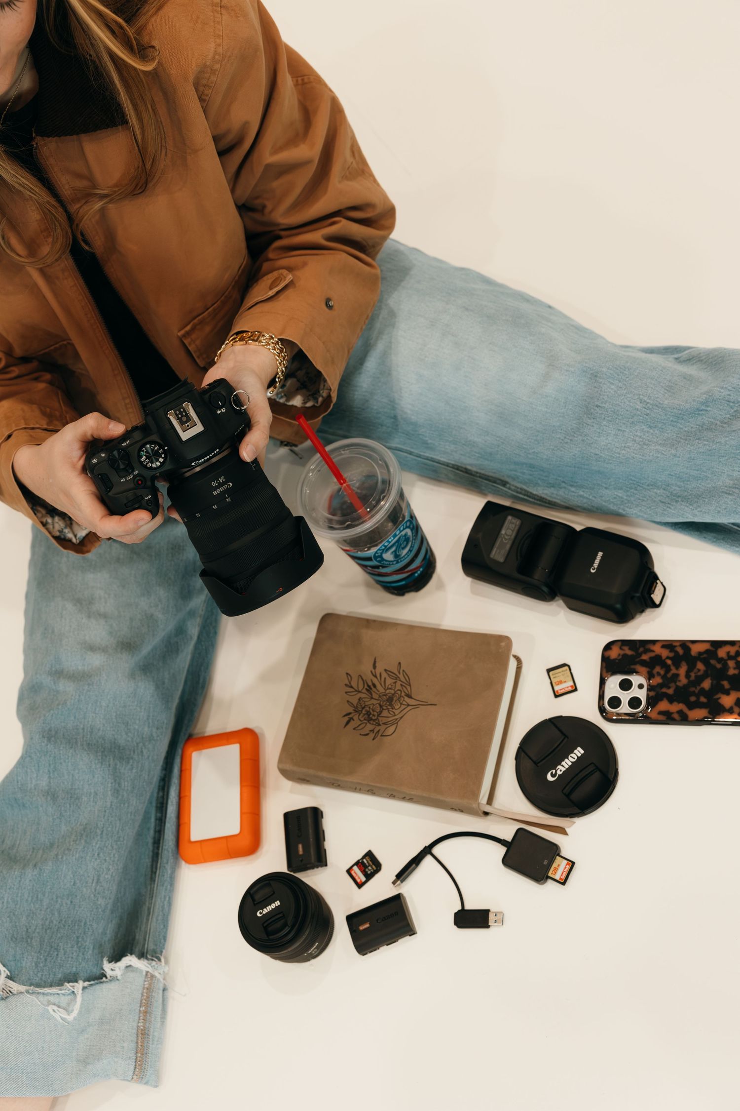 Photographer sitting with Canon camera, lens, memory cards, hard drive, notebook, and iced drink on white background.