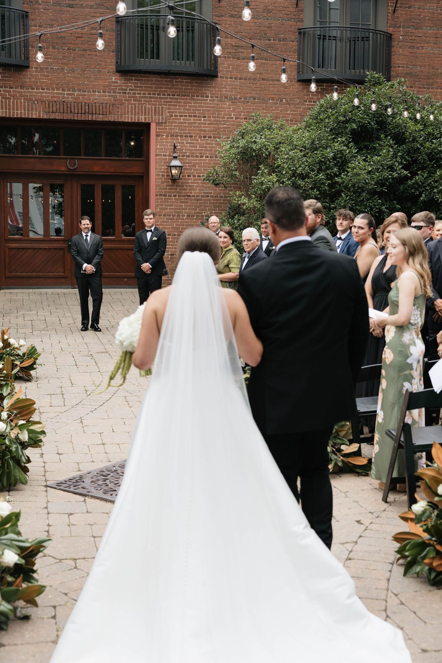 Bride in cathedral veil walks down outdoor brick courtyard aisle with escort toward waiting groom.