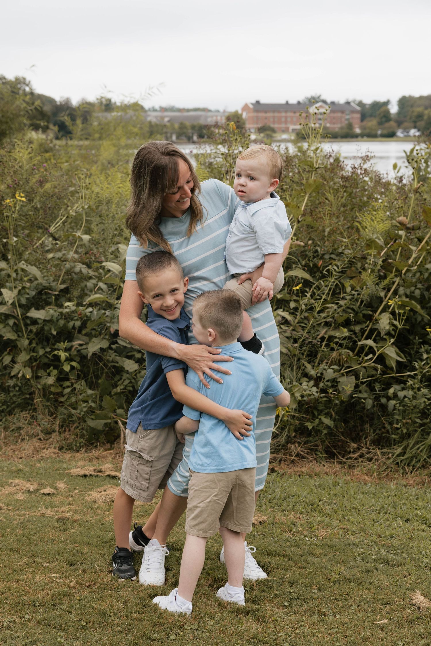 A mother holds a baby while two young boys hug her outdoors near a lake and lush greenery.