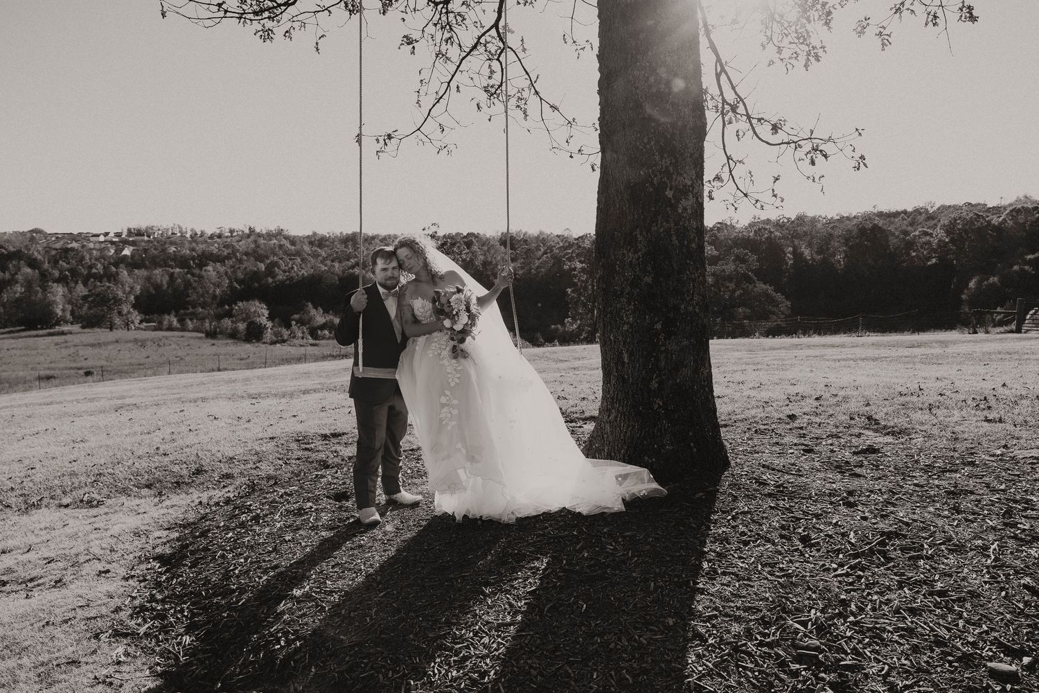 Bride and groom share intimate kiss near tree swing in sunlit black and white wedding photo.