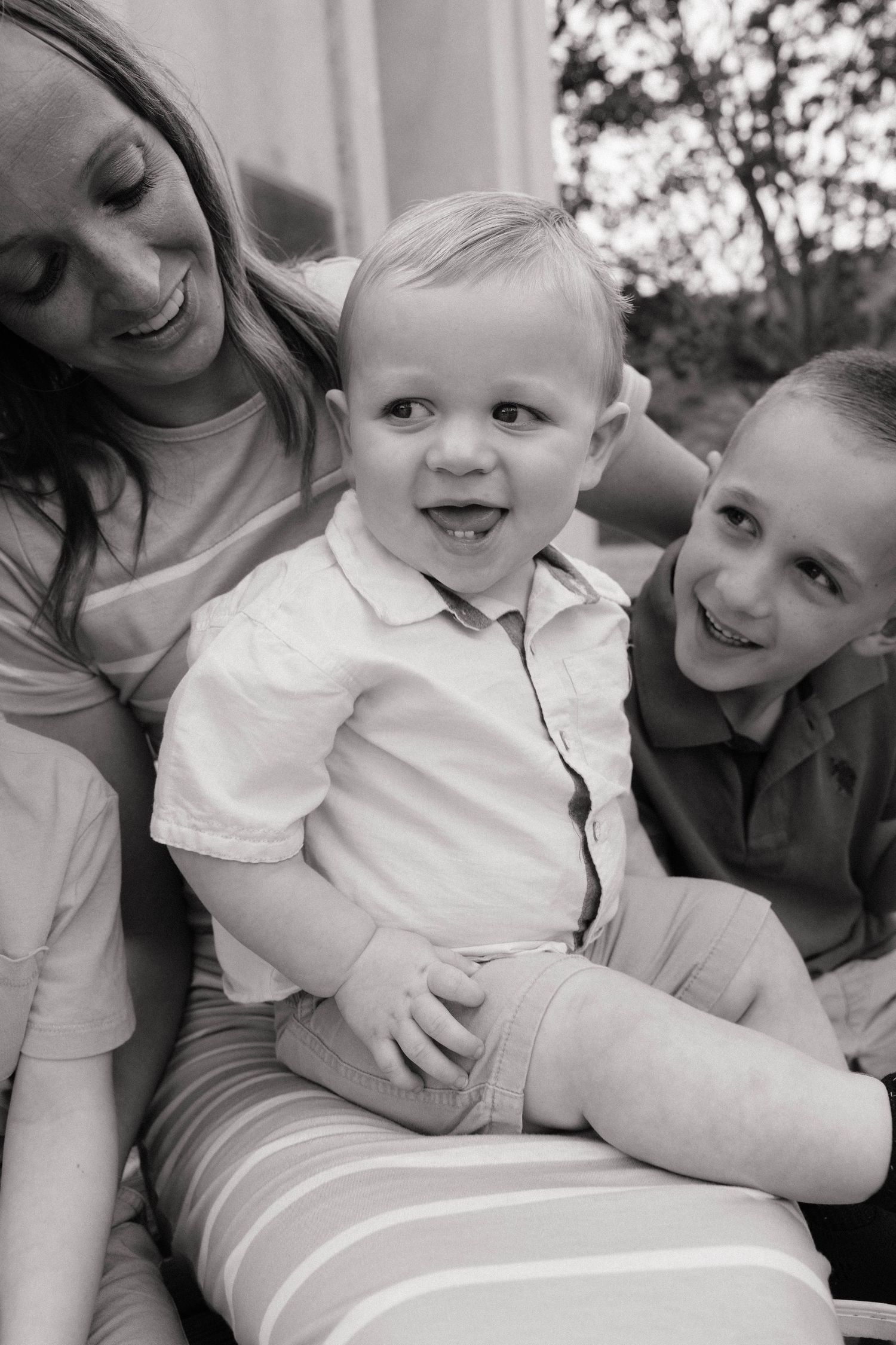 Black and white photo of a smiling mother and older boy with a happy toddler sitting on a striped blanket outdoors.