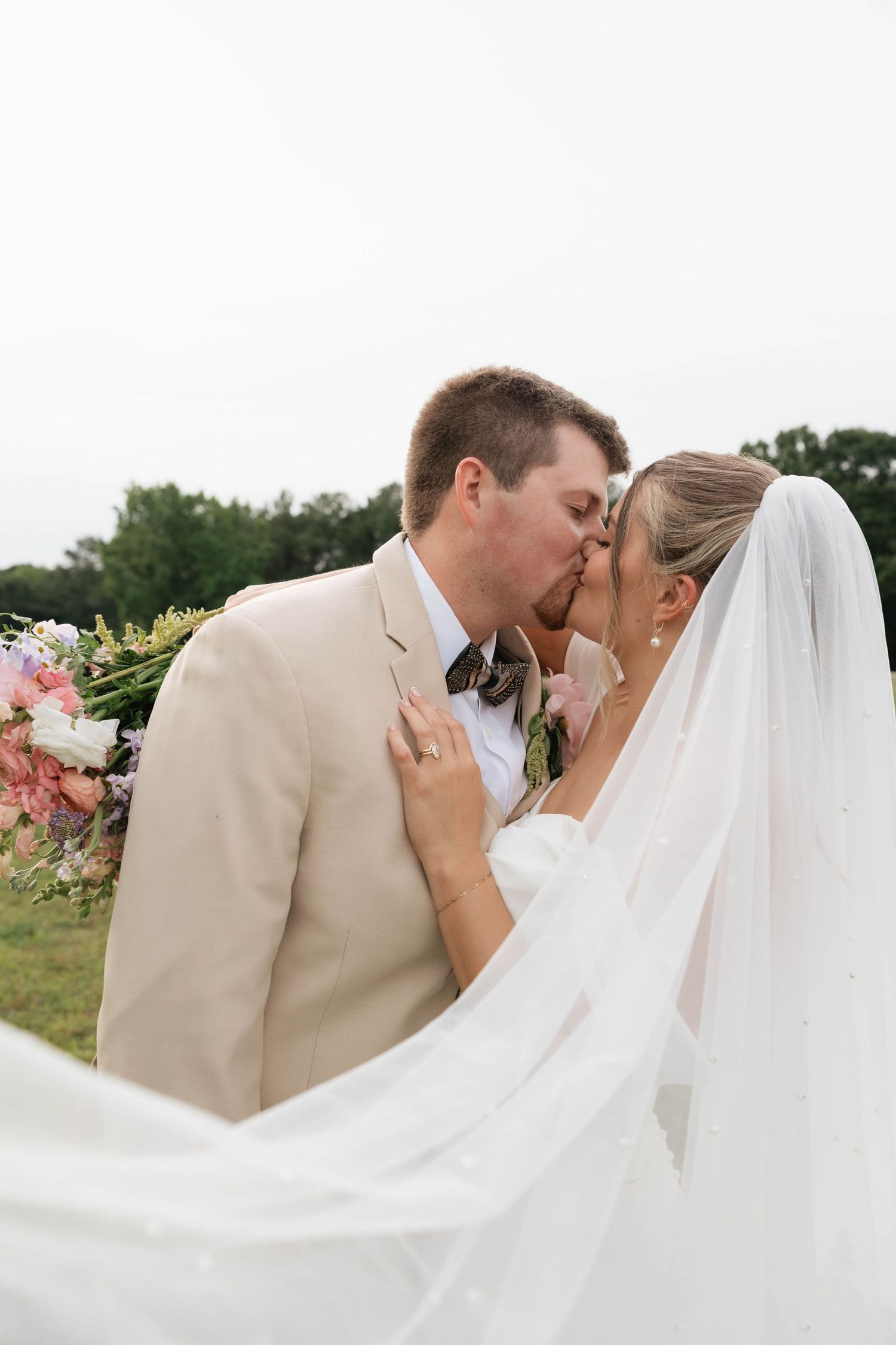 A bride and groom share a romantic kiss outdoors, with her white veil flowing and a colorful bouquet nearby.