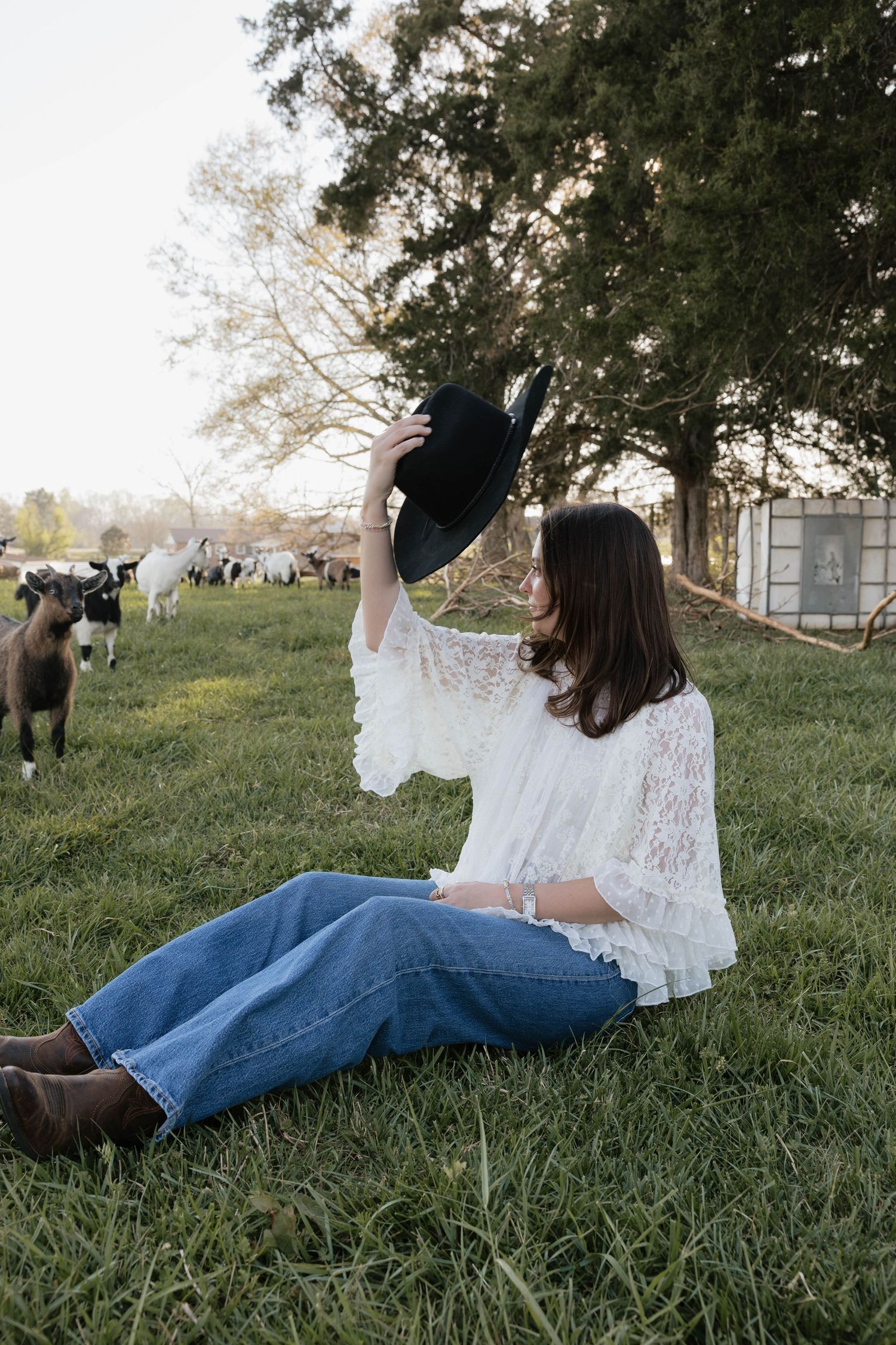 Woman in white lace top and jeans sits on grass holding black cowboy hat with goats grazing in background.