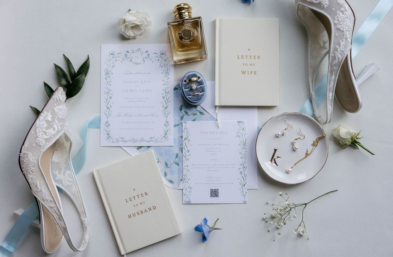 Elegant wedding flat lay with floral invitations, perfume, lace heels, love letters, and delicate blue ribbon accents.