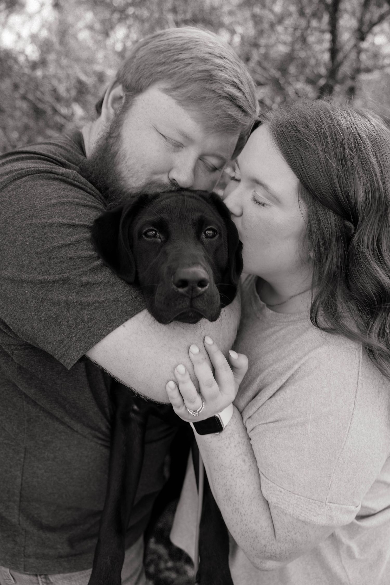 Black and white portrait of couple kissing their black Labrador puppy with engagement ring visible.