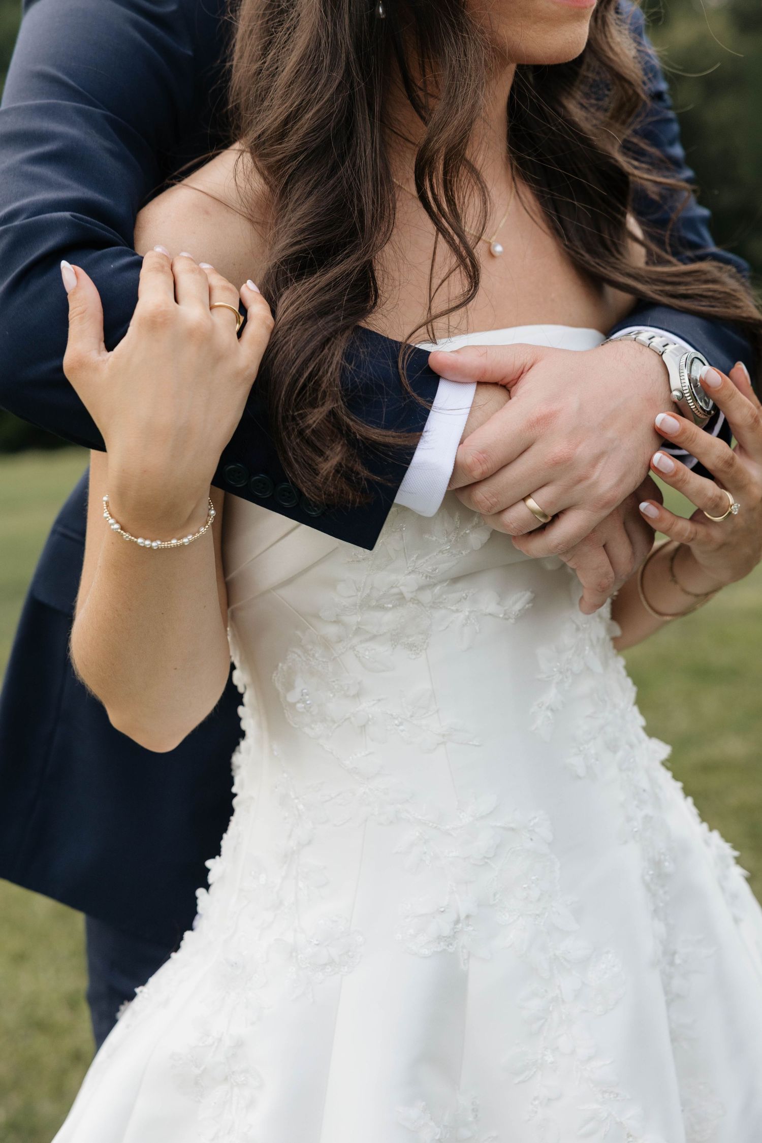 Bride in white lace gown embraces groom in navy suit, showcasing wedding rings and pearl bracelet outdoors.
