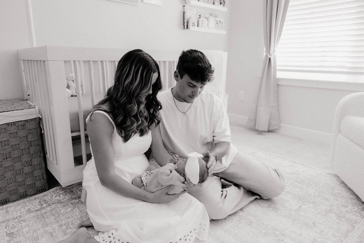 Black and white photo of new parents sitting on nursery floor, lovingly cradling their newborn baby together.