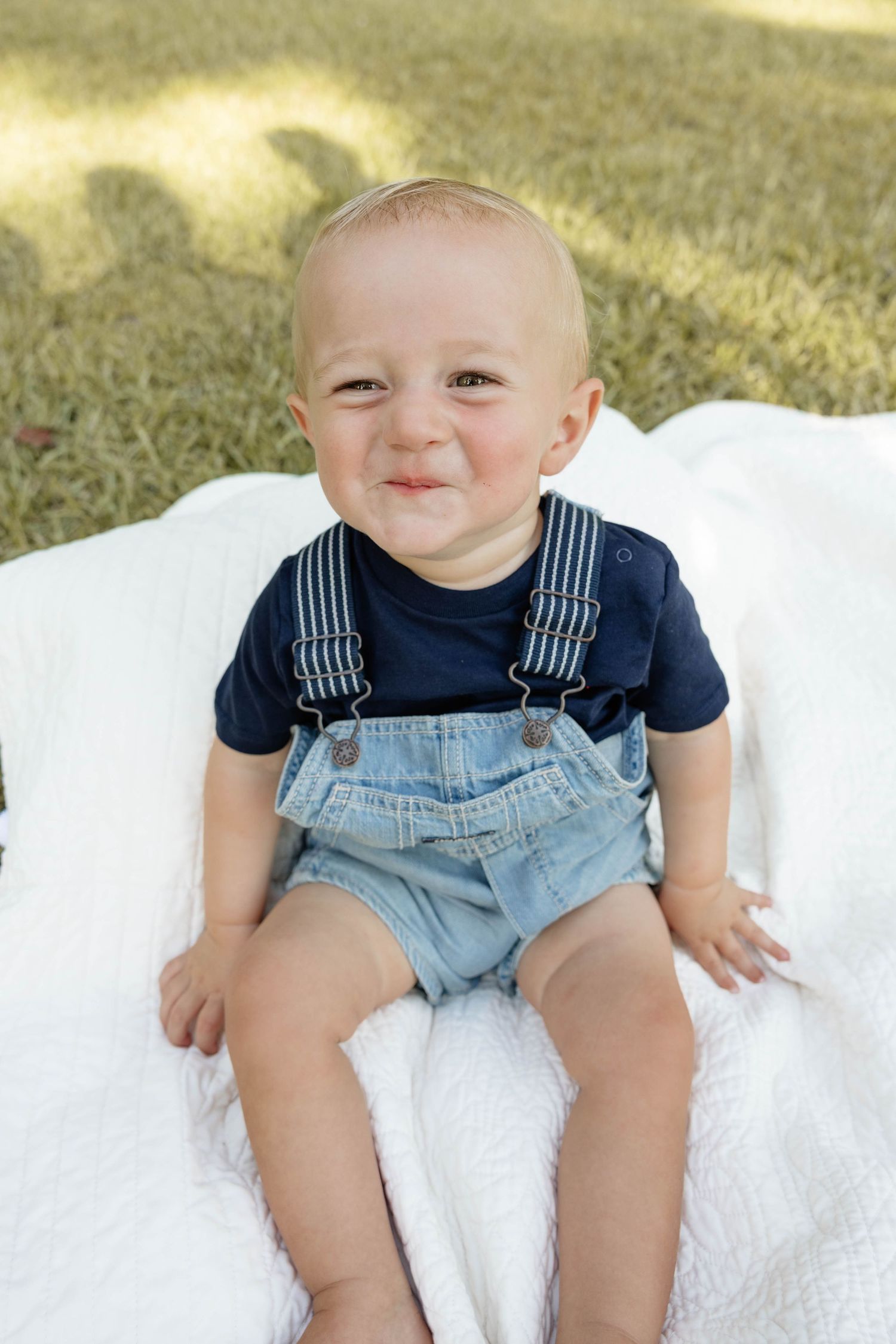 Smiling baby boy in denim overalls and navy shirt sitting on white blanket outdoors on green grass.