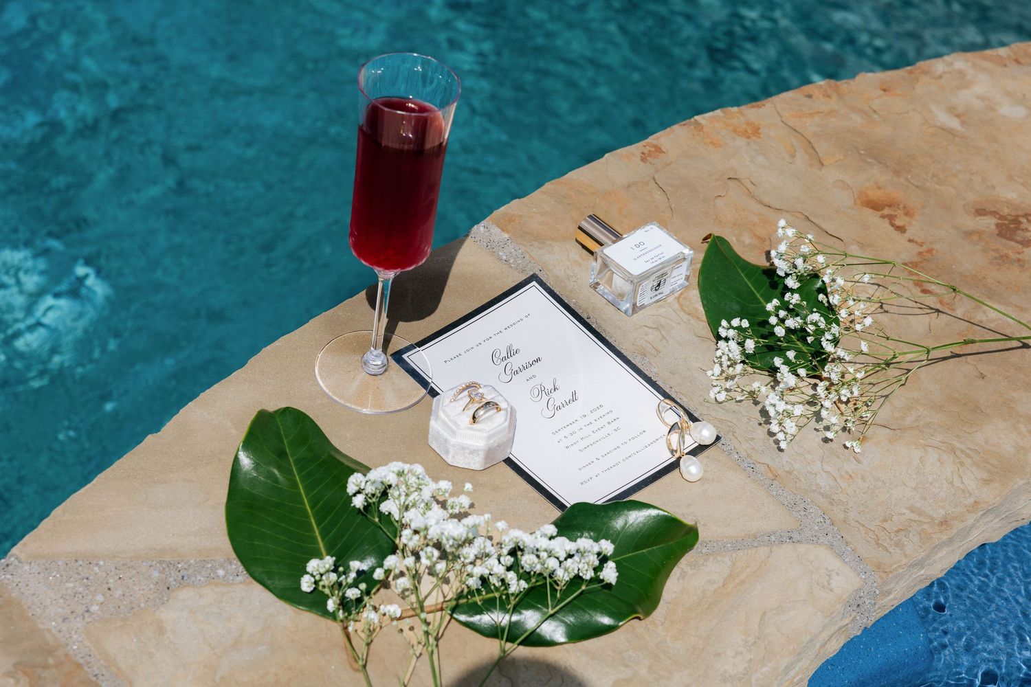 Wedding stationery display beside a red cocktail, flowers, and rings on a poolside stone ledge.