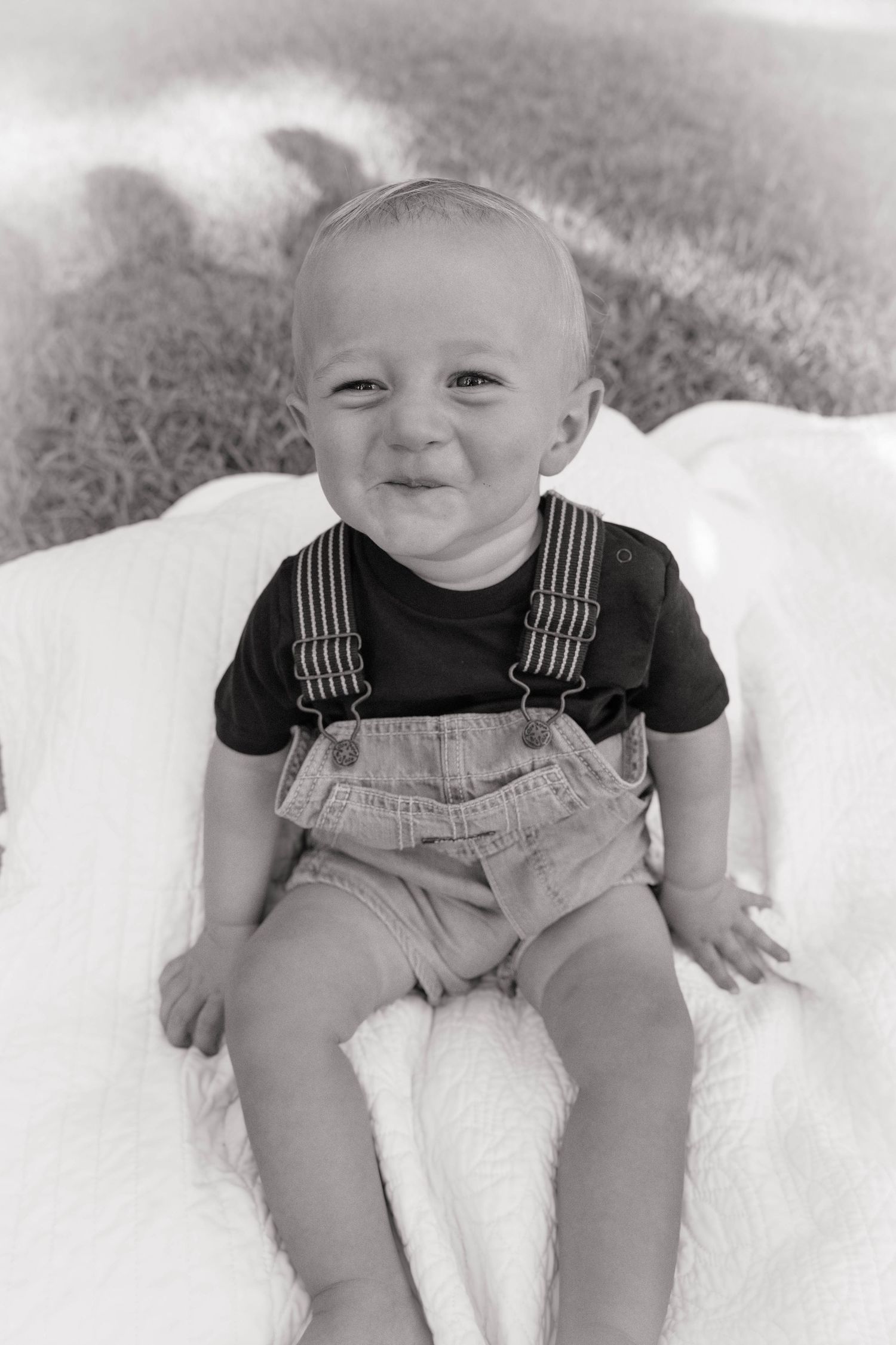 Smiling baby boy in denim overalls and black shirt sits on white blanket in black and white portrait photo.