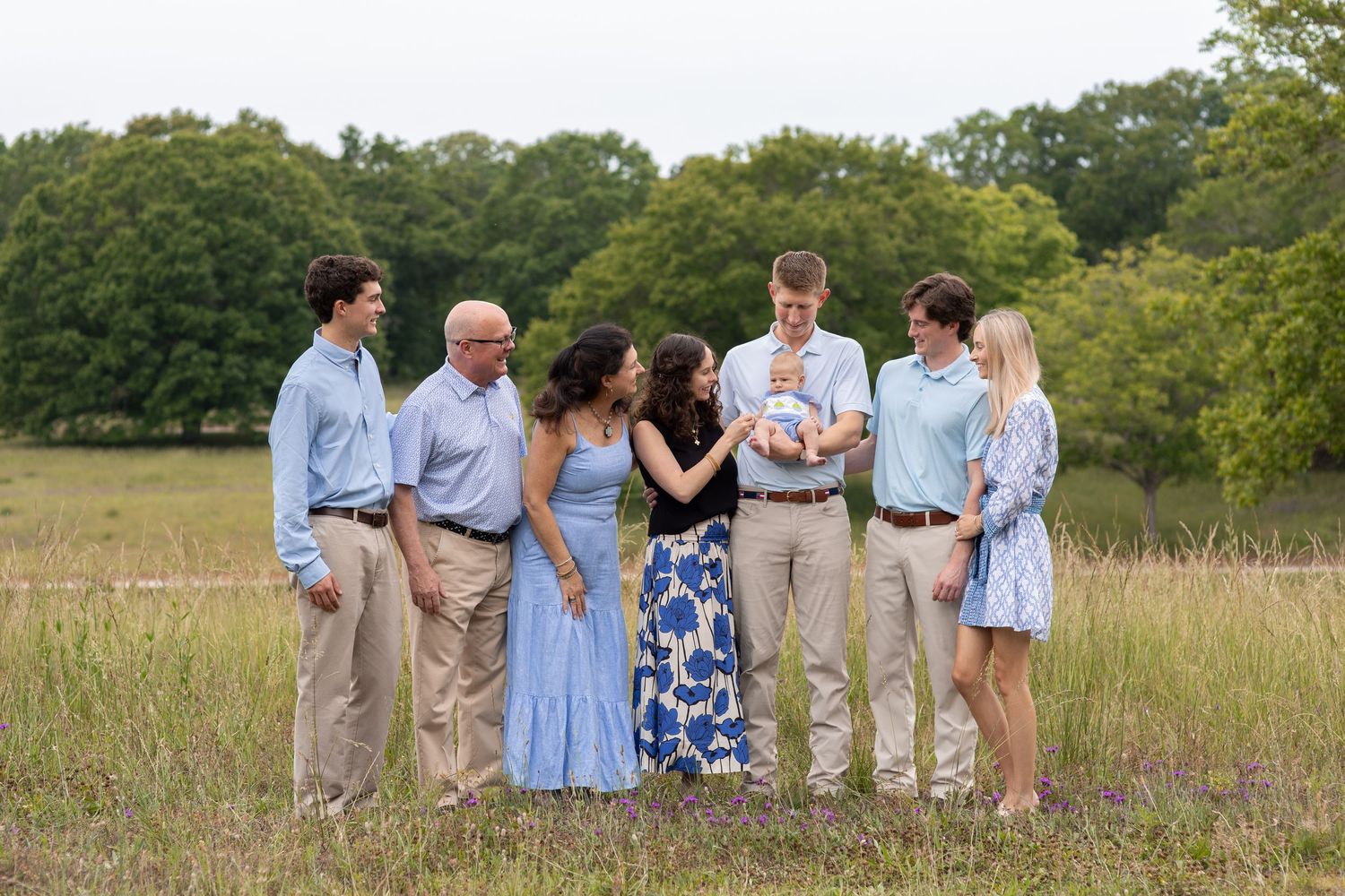 Family of seven gathered outdoors in a grassy field, dressed in blue and khaki, admiring a newborn baby.