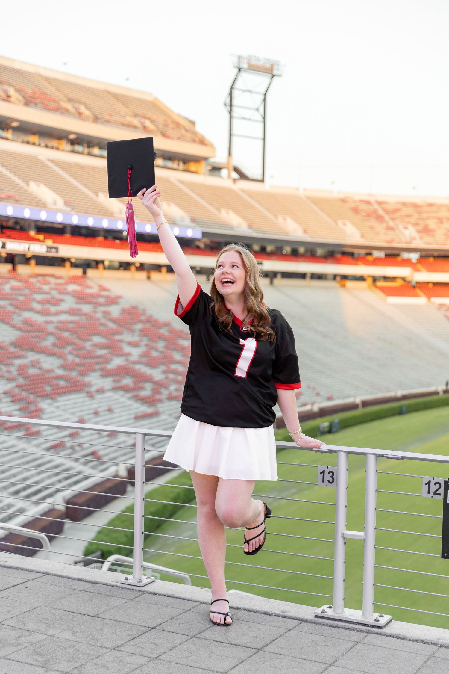 Young woman in black jersey celebrates graduation holding cap aloft in a college football stadium.