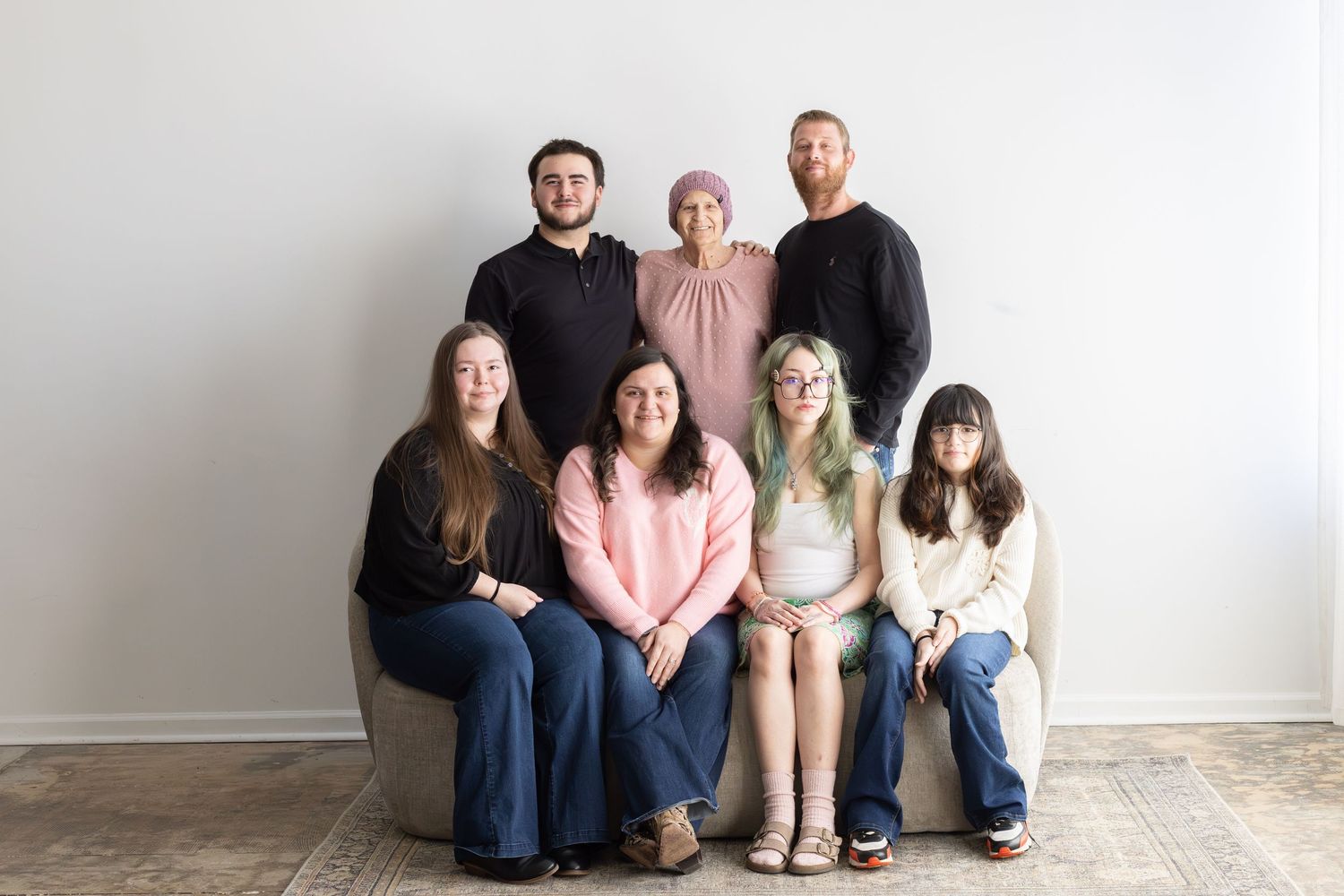 Group of seven people posing for a family portrait, three standing and four seated, in a bright minimalist studio.