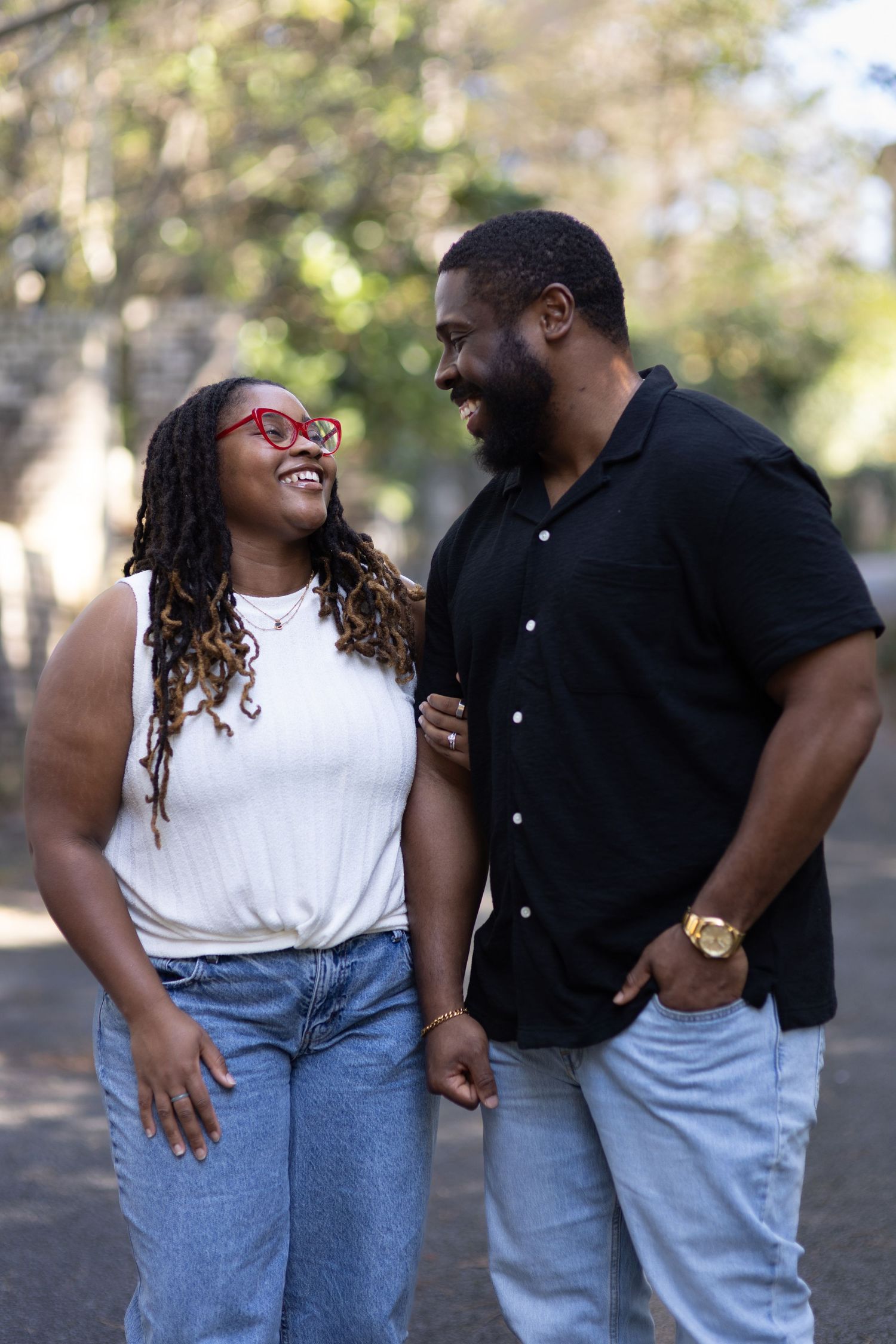 A smiling couple holds hands and gazes at each other while walking outdoors, woman wearing red glasses and white top.