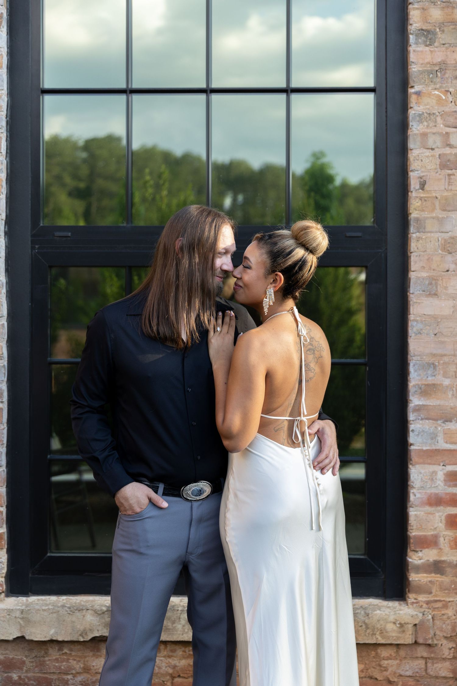 Couple sharing a romantic kiss in formal attire, white backless gown and black shirt, posed in front of a brick wall window.