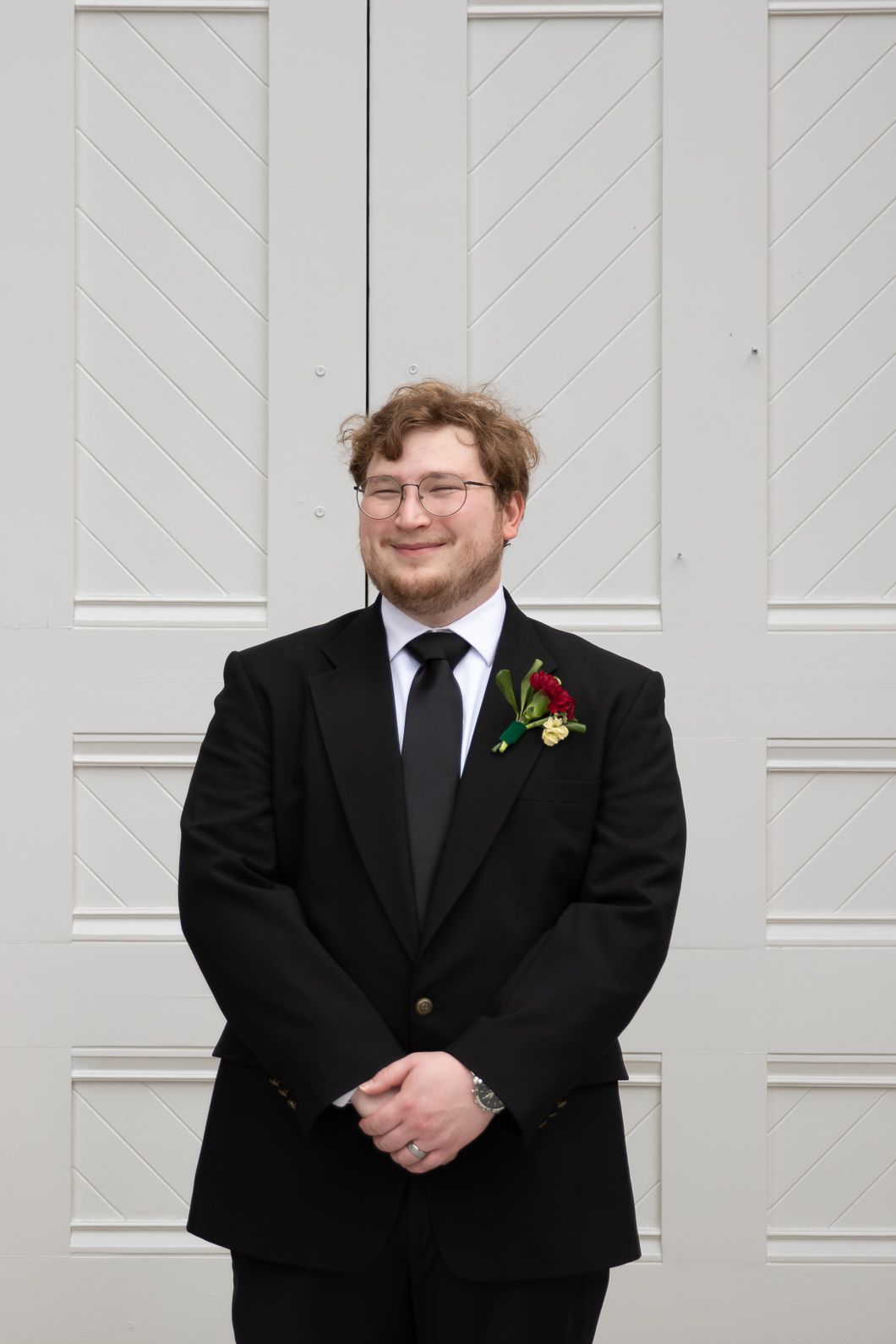 Young man in black tuxedo with red boutonniere and glasses smiling in front of white paneled door.
