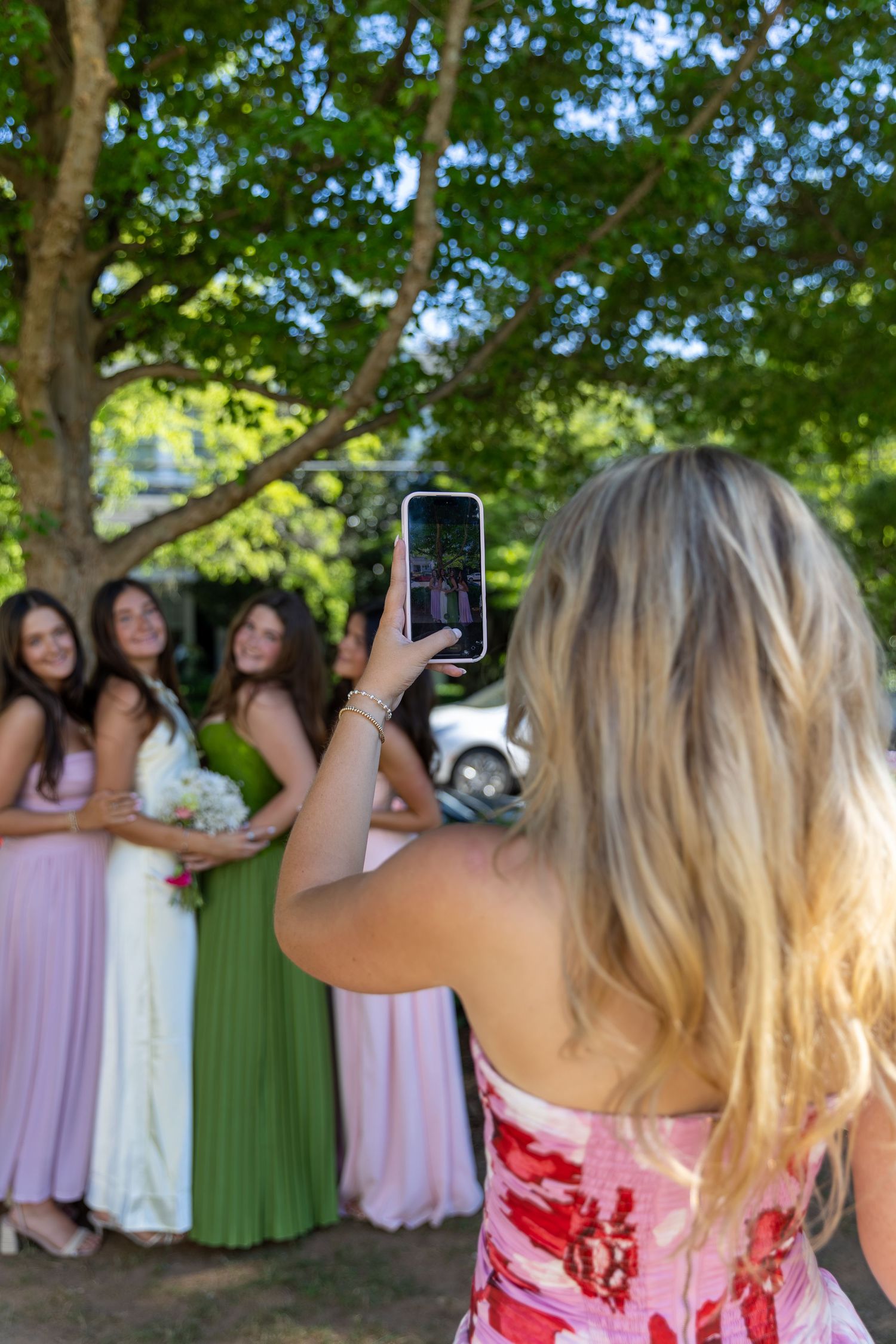 Blonde woman photographing bridesmaids in colorful gowns outdoors under a tree using a smartphone.