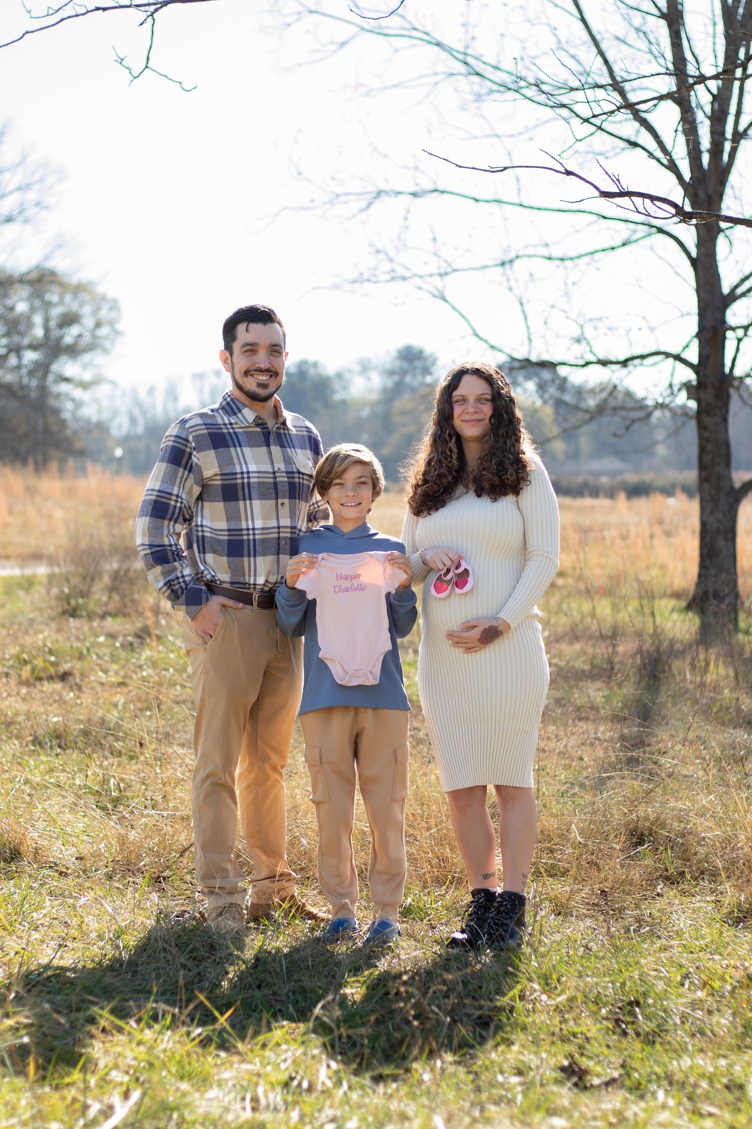 Pregnant woman in white dress stands outdoors with husband and son holding pink baby onesie for gender reveal photo.