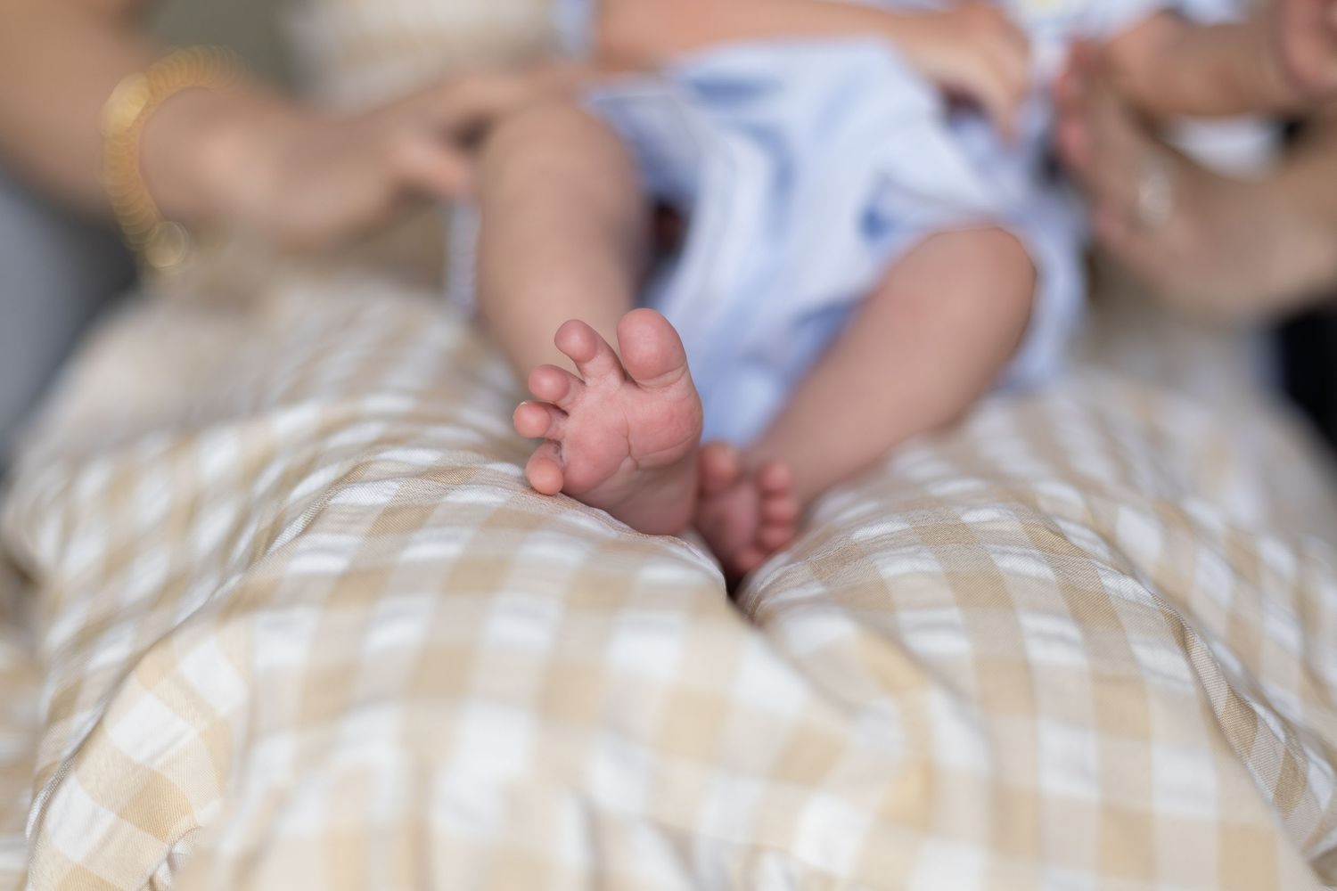 Close-up of tiny newborn baby feet resting on a soft gingham blanket, with adult hands visible in background.
