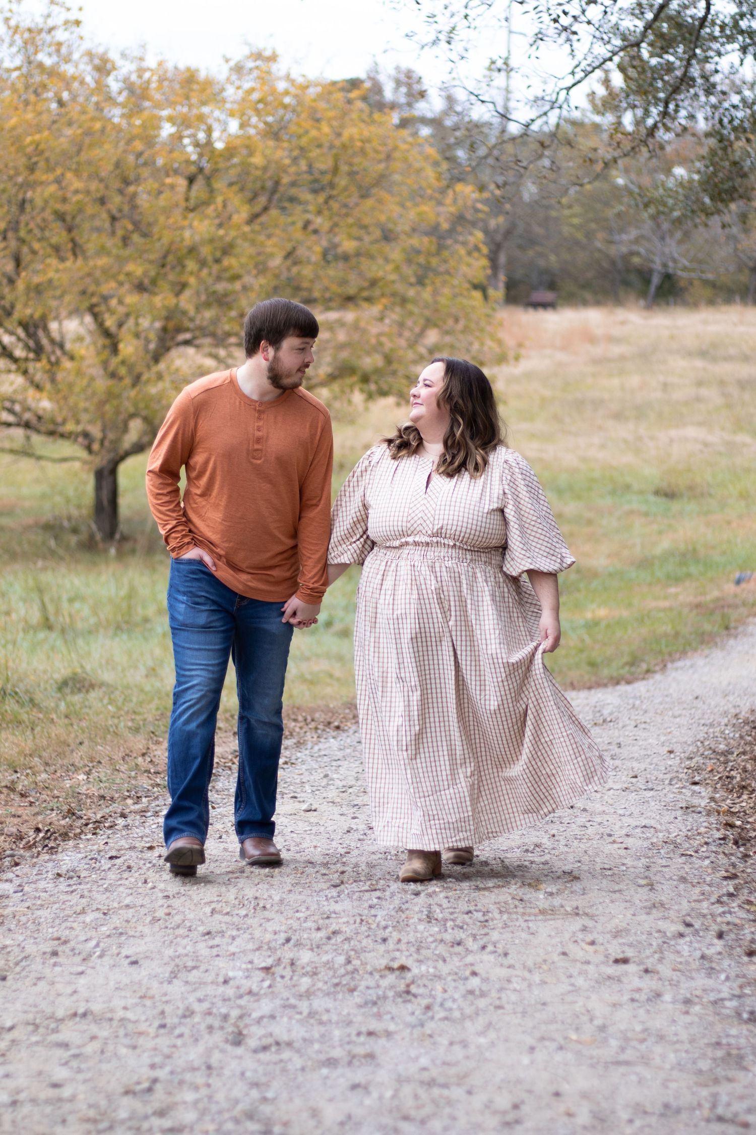 Couple holding hands walking on a gravel path surrounded by autumn trees in a rural setting.