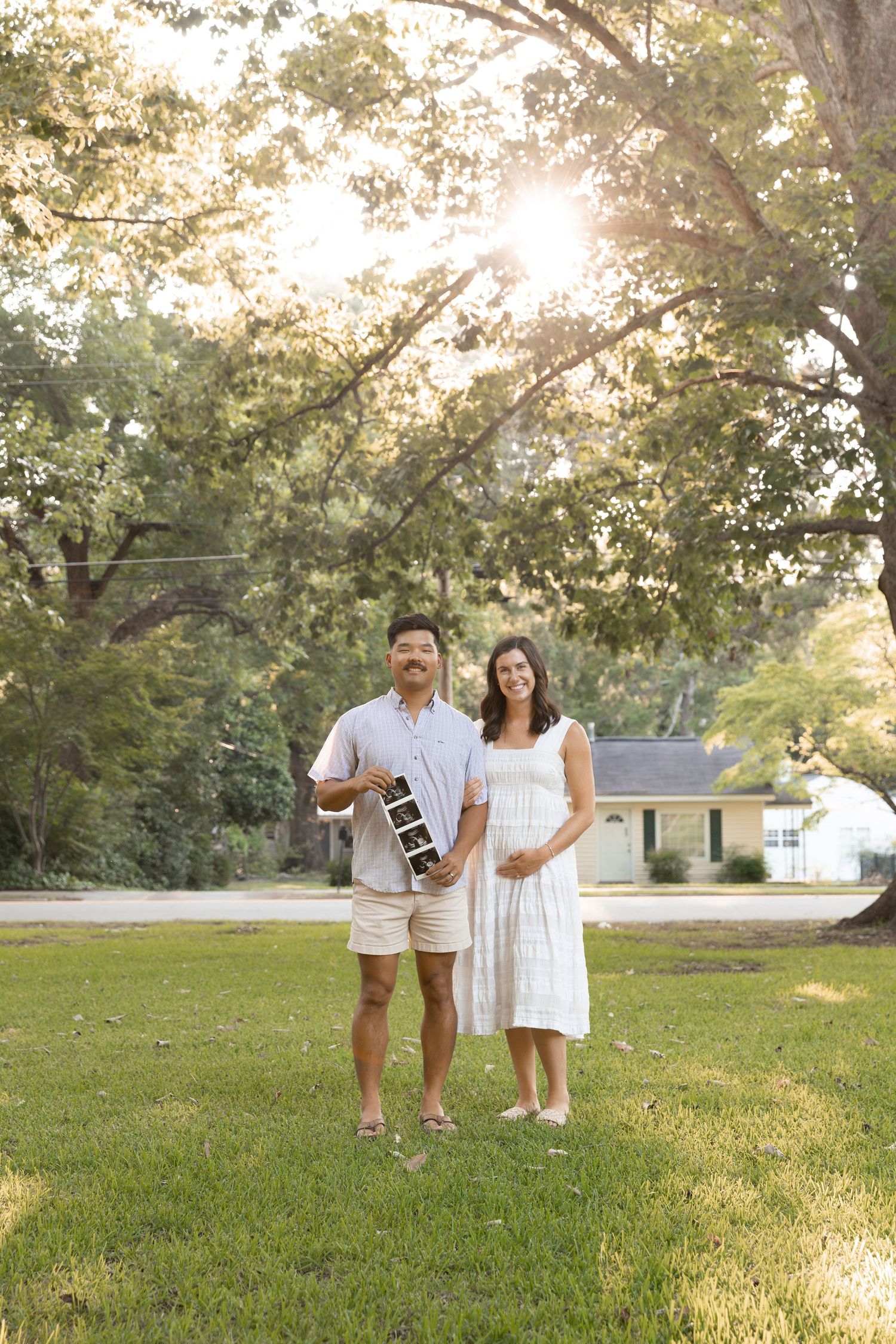 A couple stands on a green lawn holding an ultrasound photo, announcing their pregnancy outdoors.