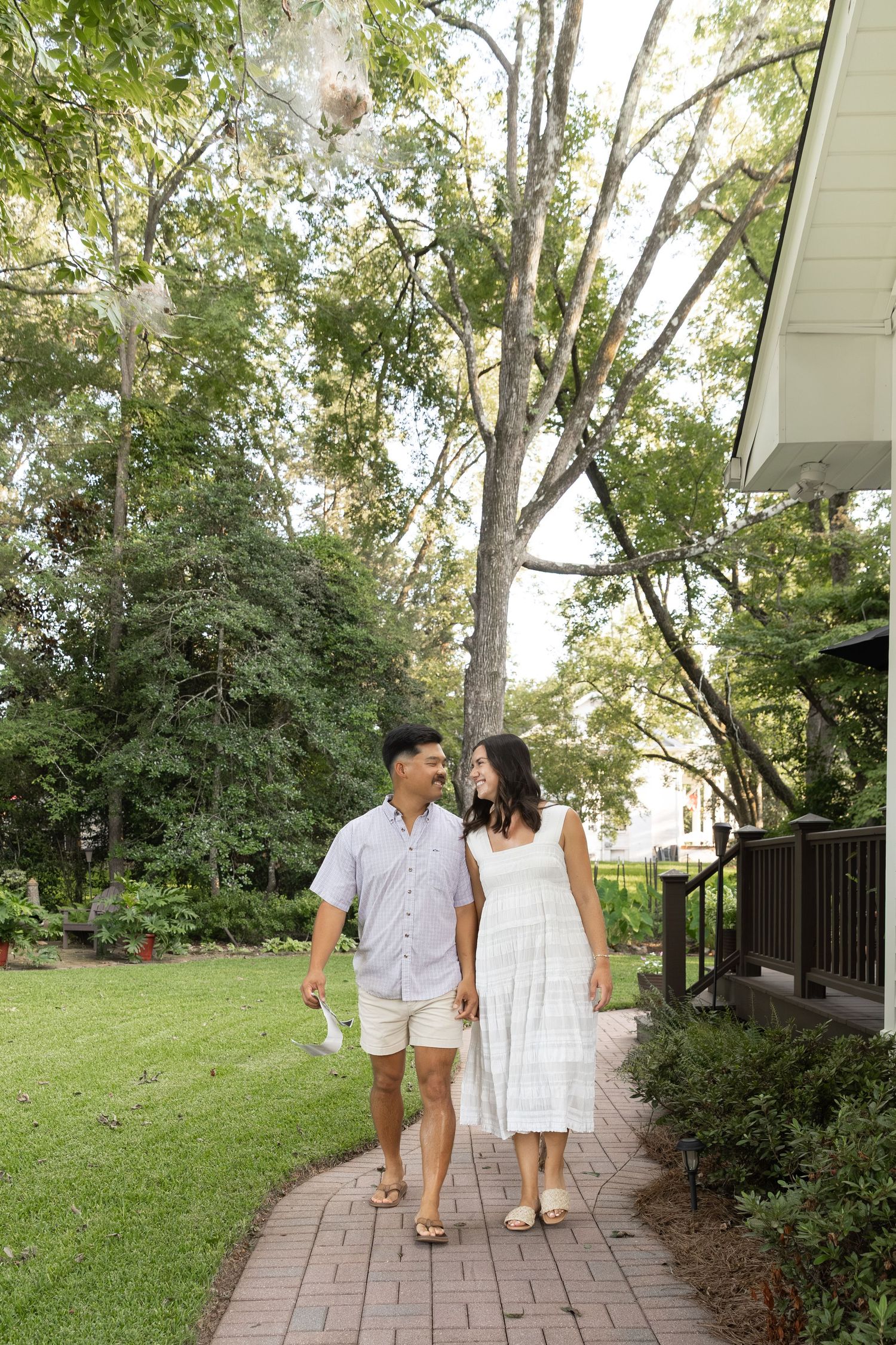 Happy couple walking hand-in-hand on brick pathway surrounded by lush green trees near a home.