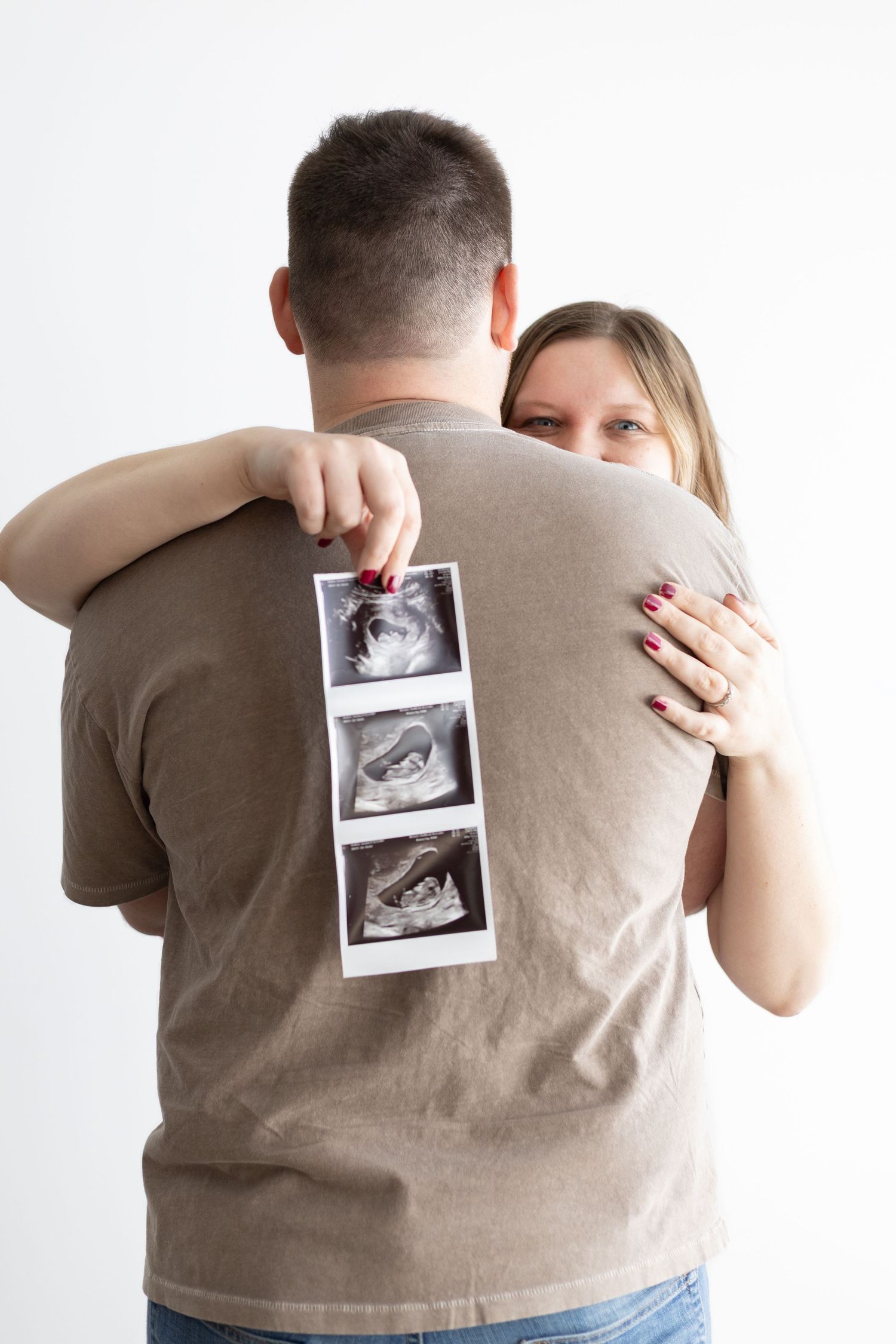 Couple embracing and holding ultrasound photos behind man's back, announcing pregnancy on white background.