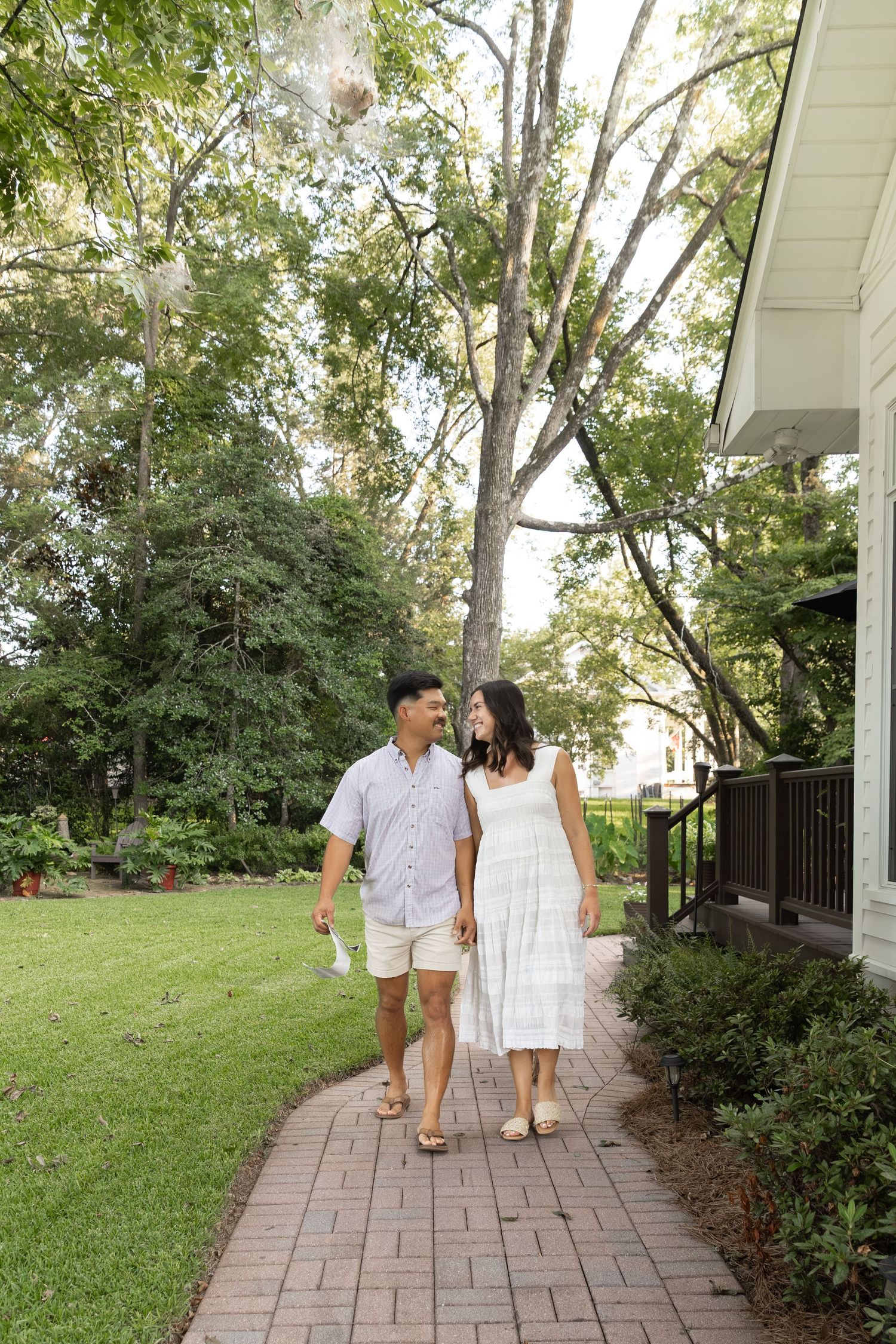 Couple walking hand in hand on a brick path beside a white home surrounded by lush green trees.