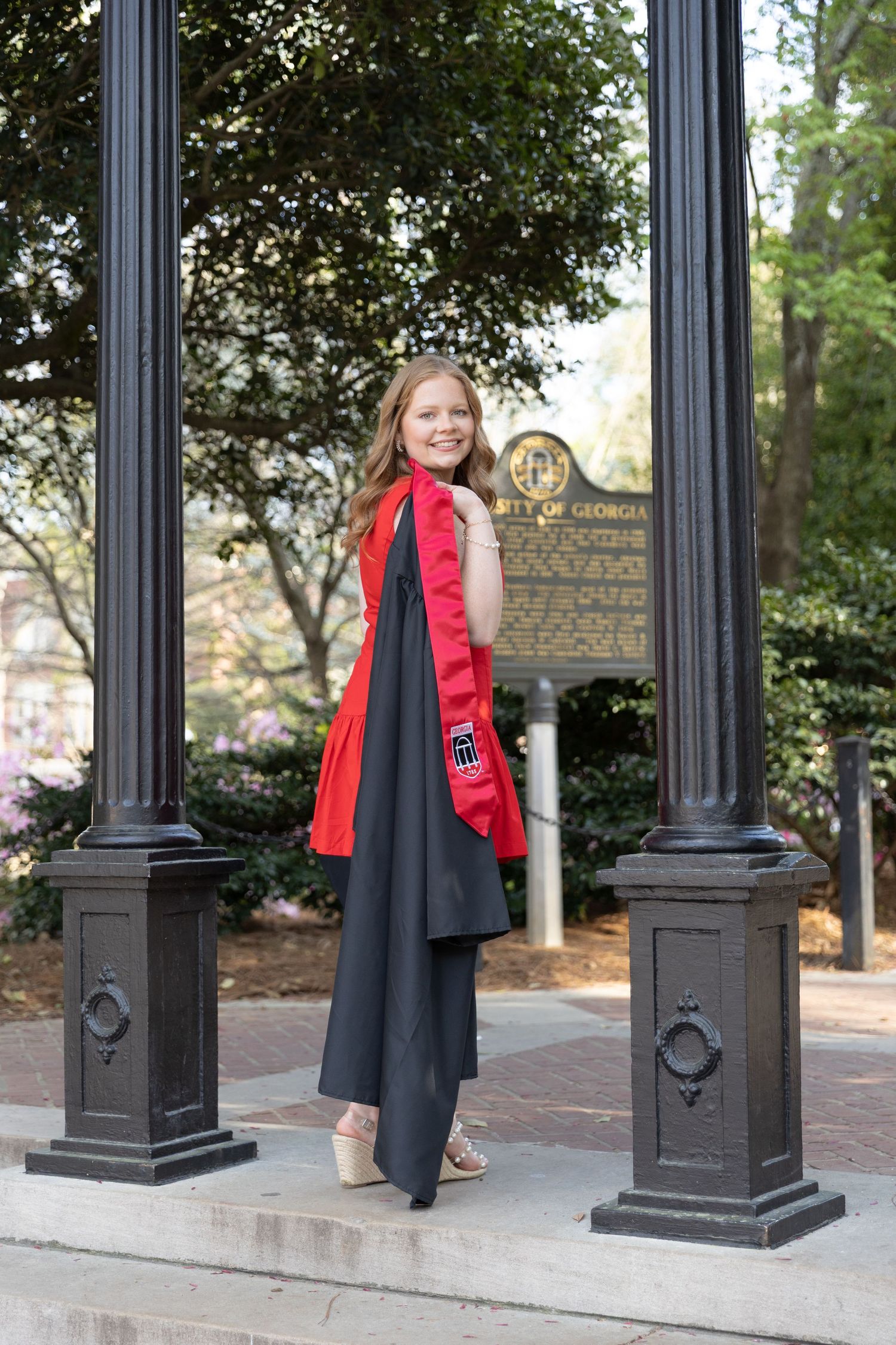 Young woman in black graduation gown with red stole poses near University of Georgia historical marker.