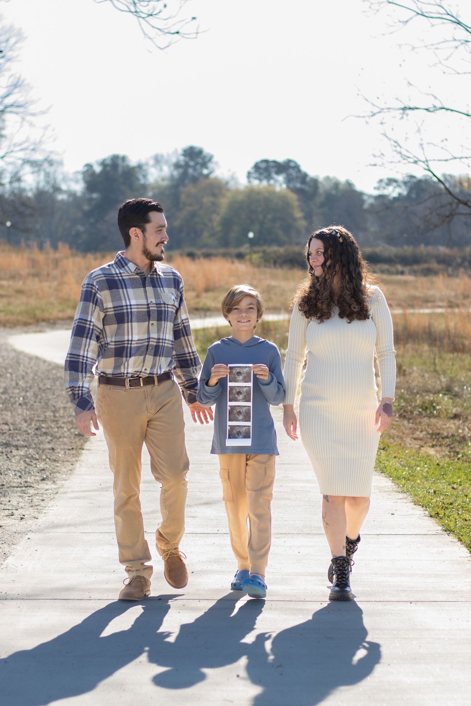 Family of three walking on a path, boy holding ultrasound photos, pregnancy announcement outdoors.