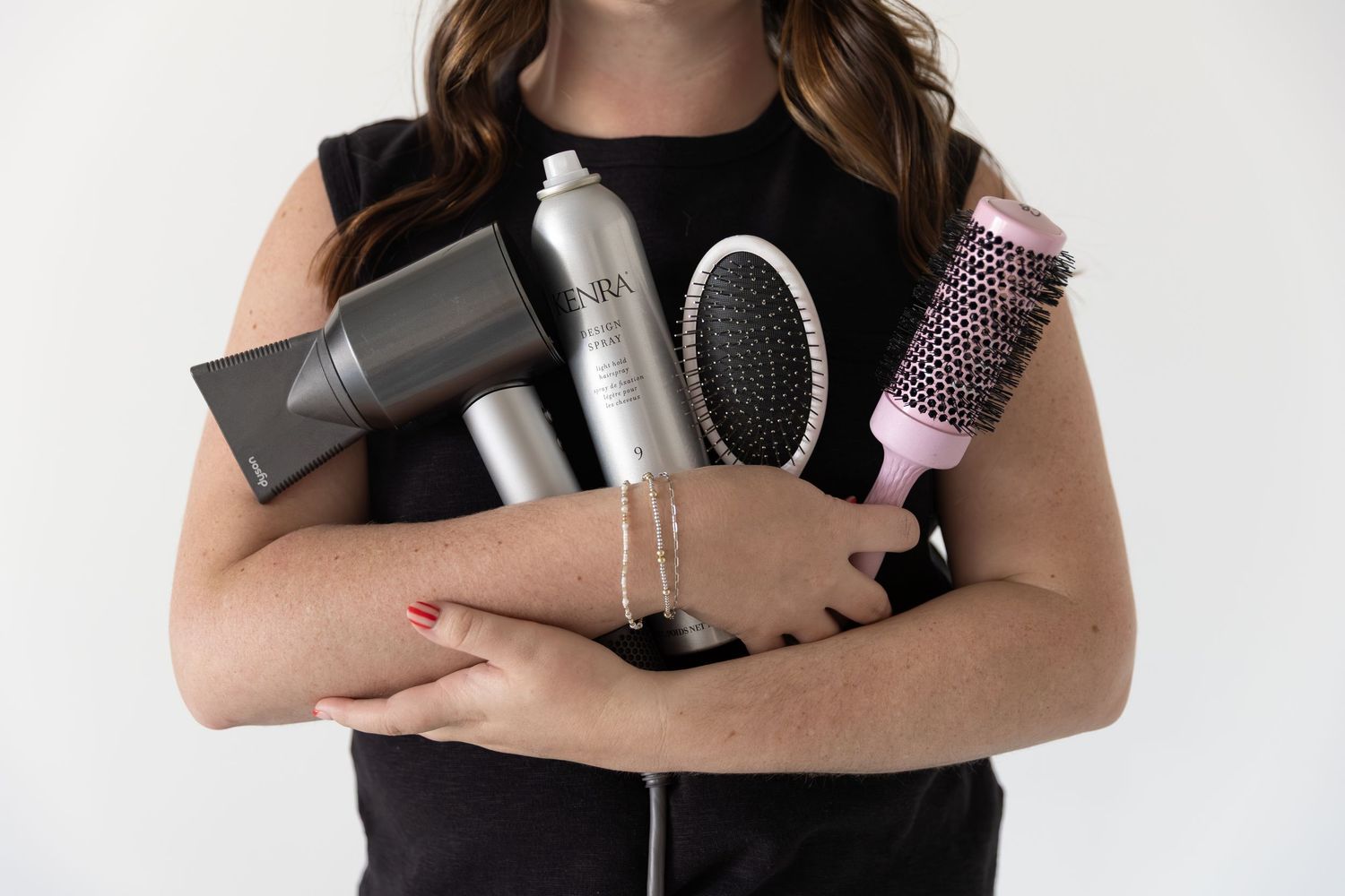 Woman holding hair styling tools including a blow dryer, hairspray, round brush, and paddle brush against white background.