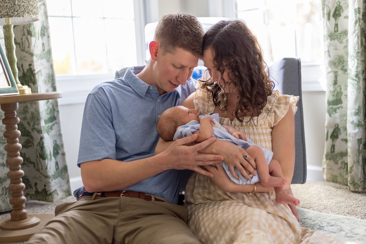 A couple sits together lovingly cradling their newborn baby in a bright, cozy nursery room.